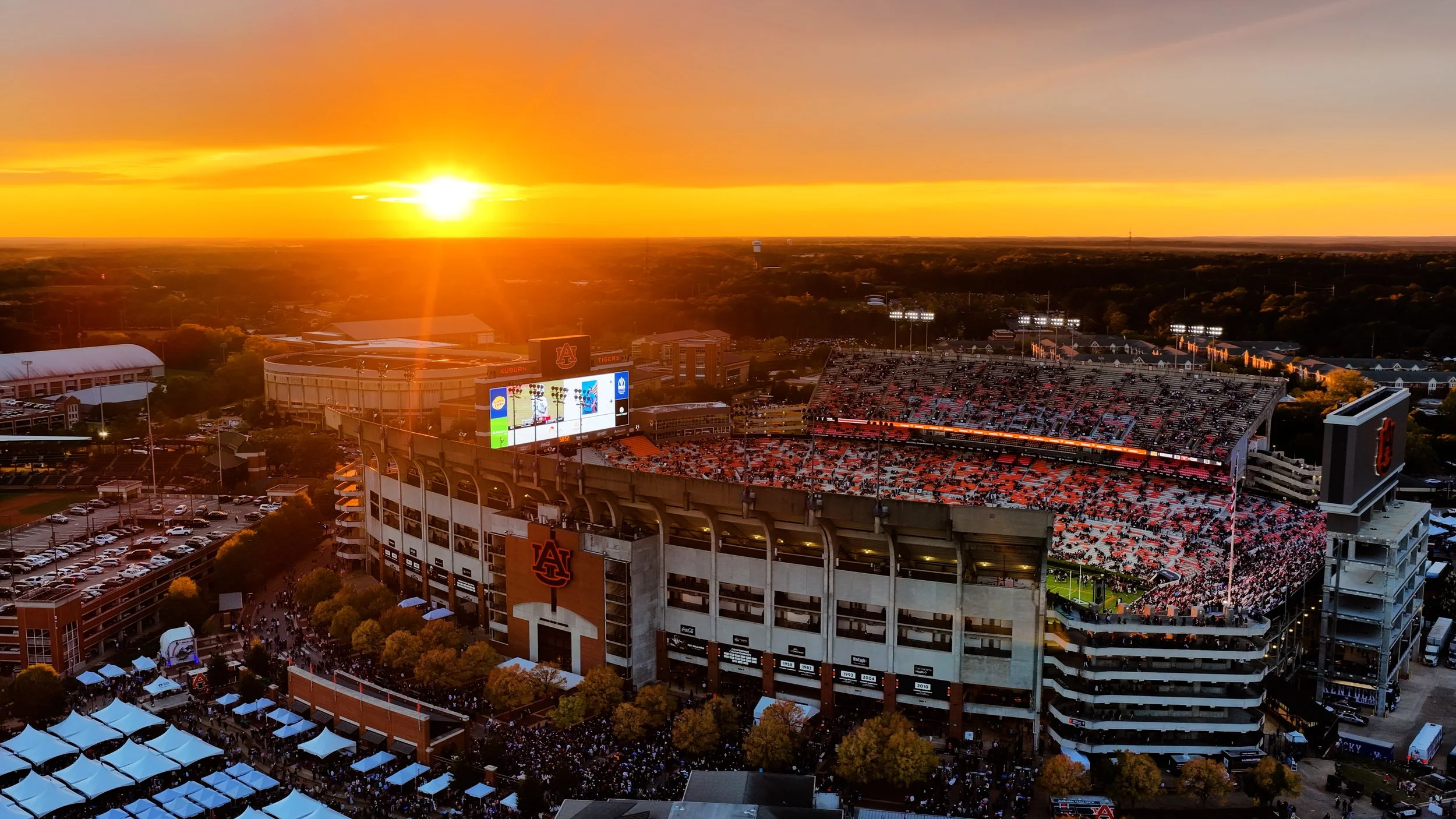 Aerial view of Auburn University football stadium at sunset, filled with fans and illuminated field, with surrounding campus and parking lot.