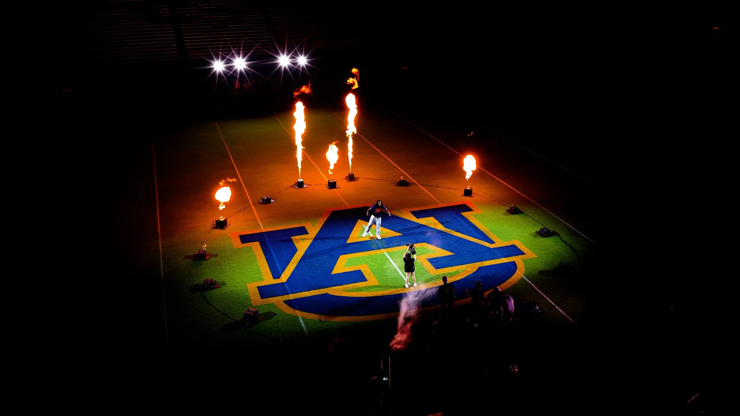 A football field with a large logo in the center, illuminated by bright spotlights. A person is standing on the logo, with smaller flames on the field around them and large flames erupting from equipment. Several bright lights are visible above, creating starburst effects.