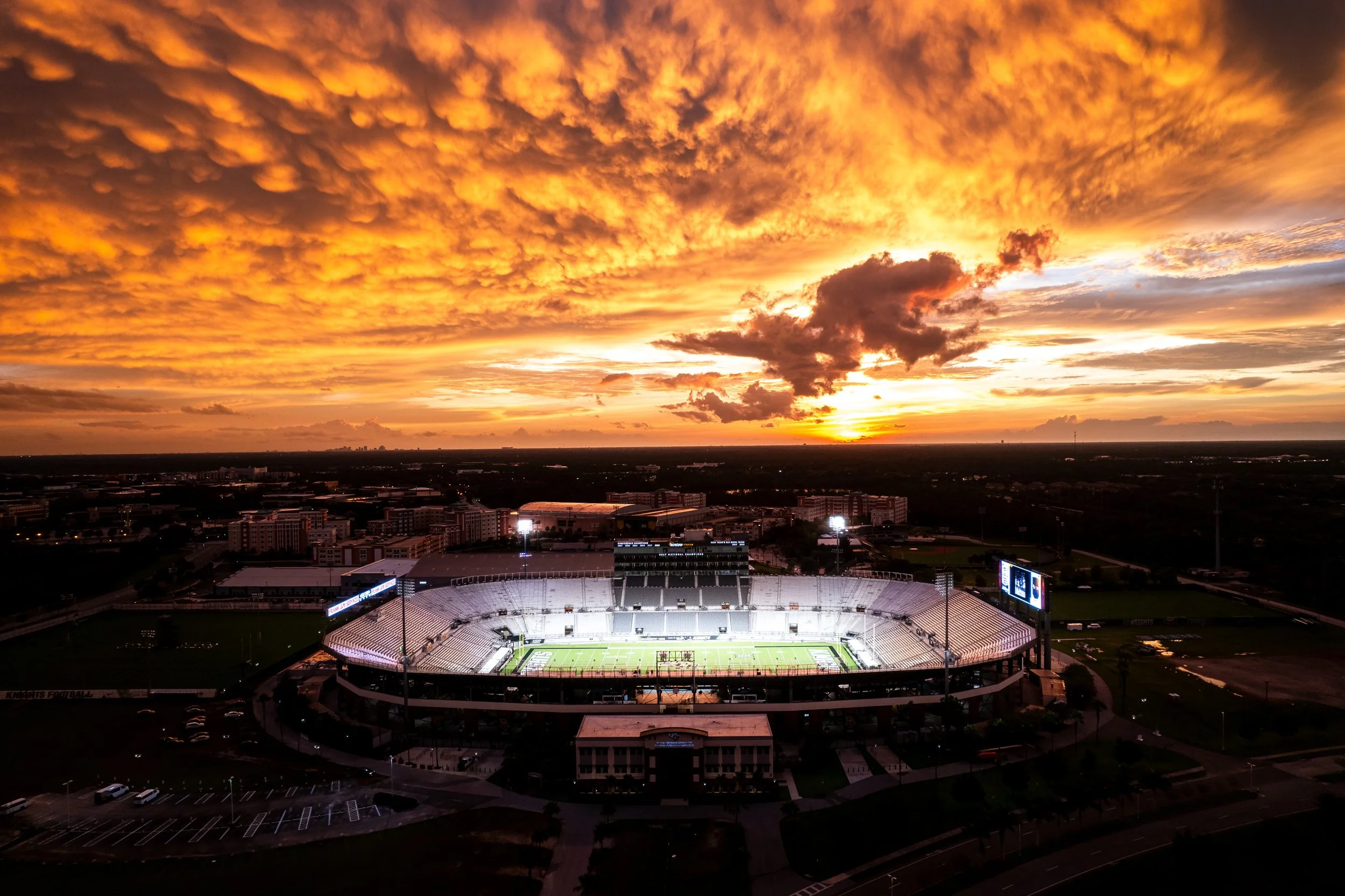 A stadium during sunset with dramatic orange and yellow clouds in the sky, cityscape in the background, and the stadium lit up for an event.