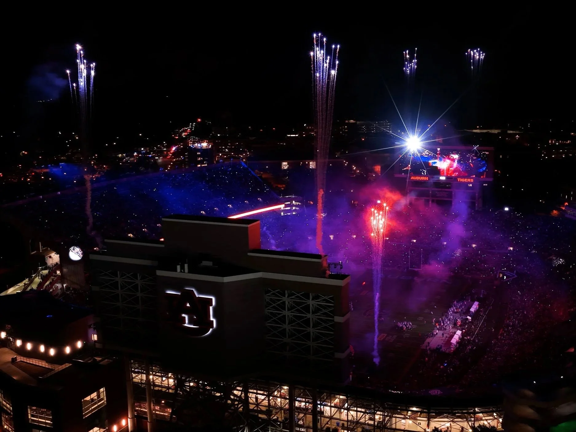 Fireworks explode over a stadium at night, with purple and white streaks lighting up the sky. There is a large illuminated sign for Auburn Tigers visible in the foreground.