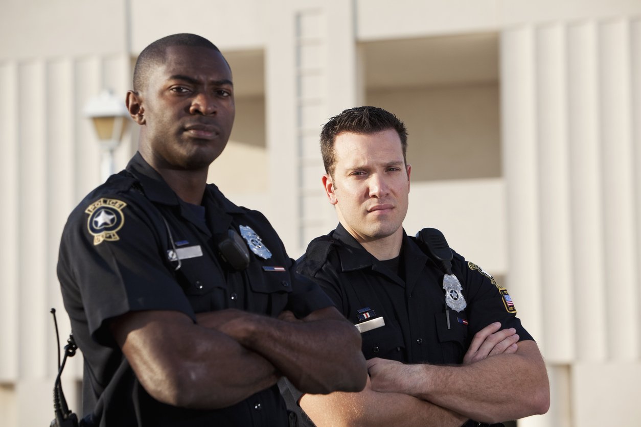 Two police officers standing outdoors with arms crossed, looking serious, in front of a building with white columns.