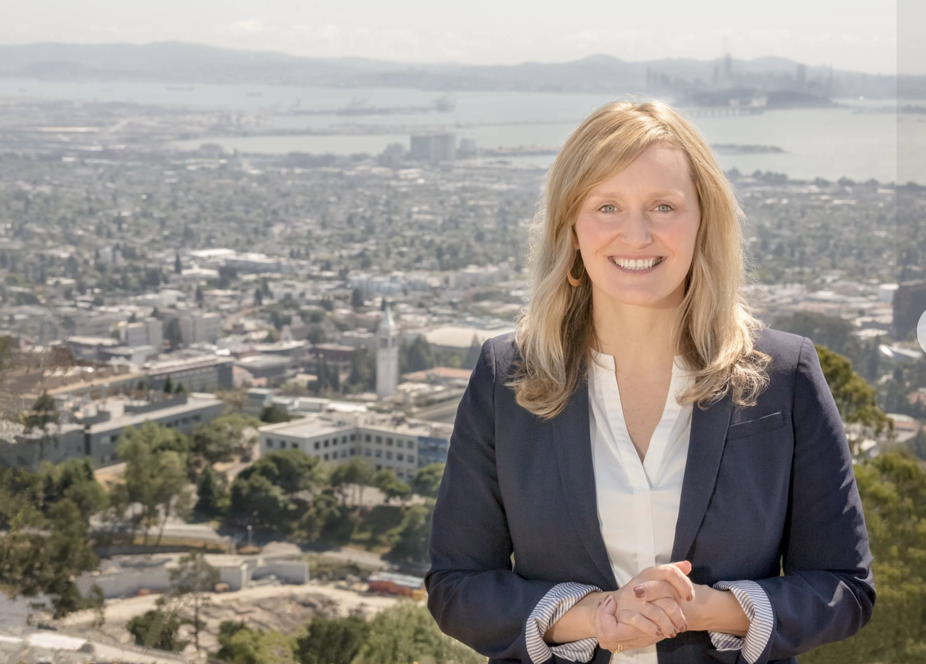 A woman with blonde hair smiling, wearing a dark blazer and white shirt, standing outdoors on a hill with a cityscape, water, and distant mountains in the background.
