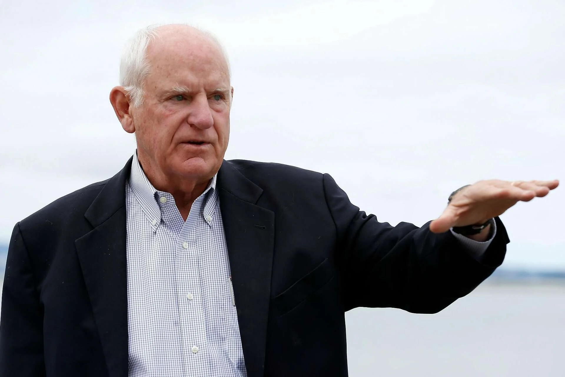 An older man with white hair wearing a dark suit jacket and a checkered dress shirt, gesturing with his right hand against an overcast sky background.
