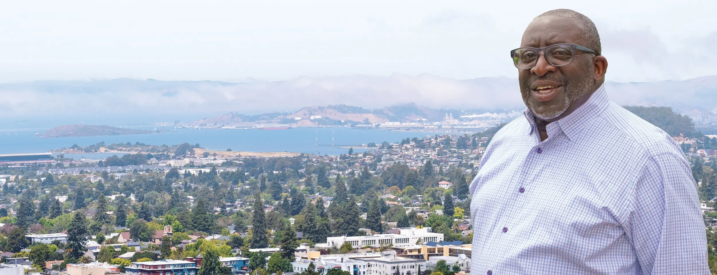 An older man with glasses and a gray beard wearing a light purple checkered shirt, outdoors with a cityscape, trees, water, and mountains in the background.