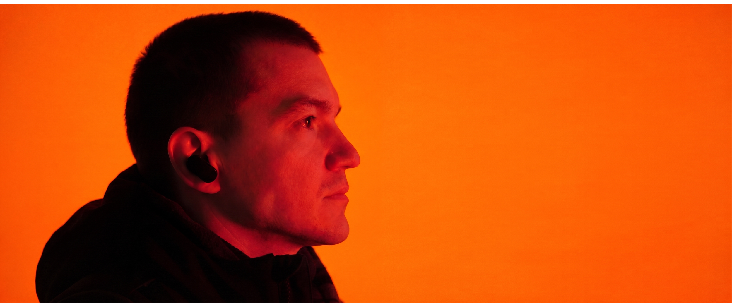 A man with short dark hair and earbud headphones, looking to the right, against an orange background.