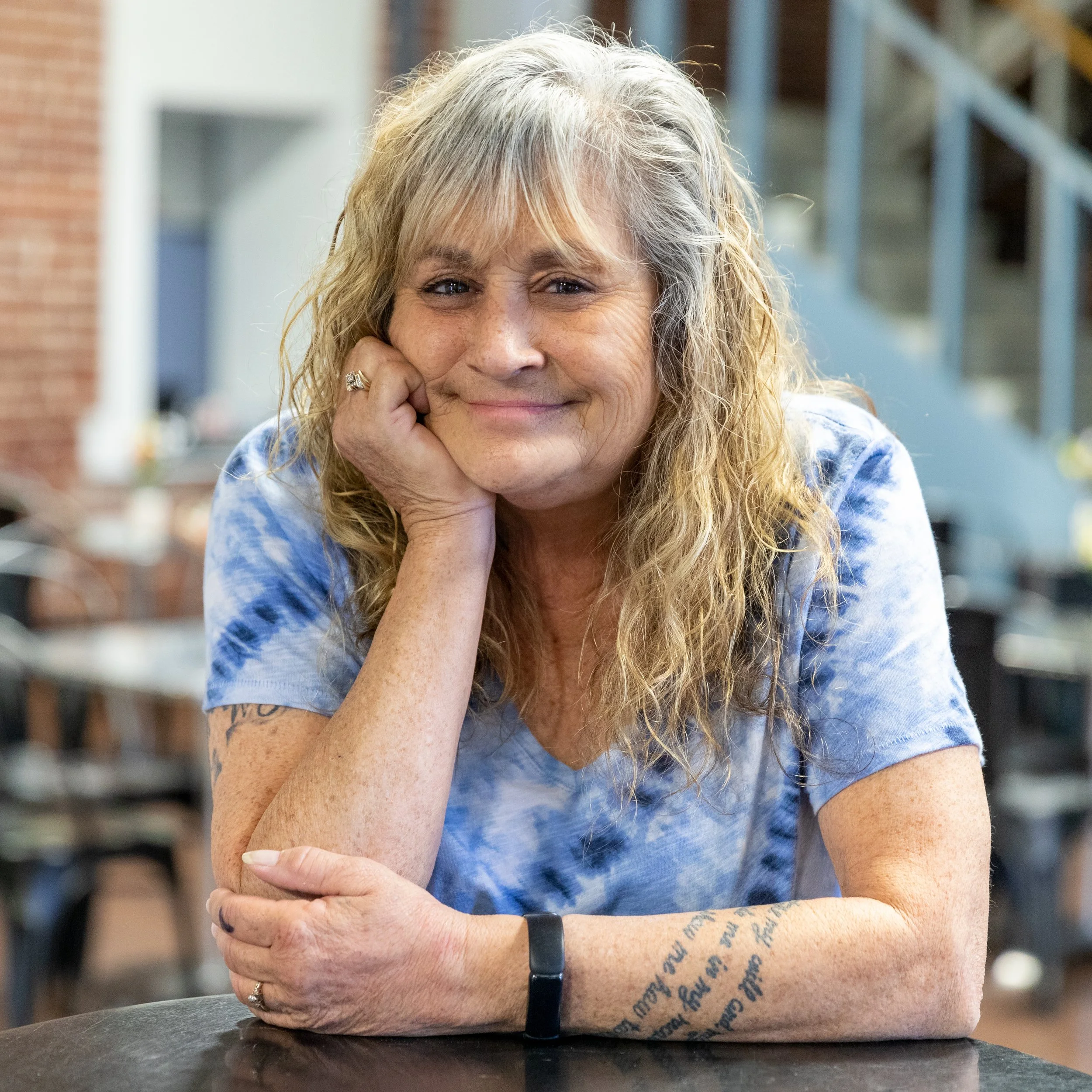 A smiling middle-aged woman with long curly gray hair, wearing a blue tie-dye shirt, resting her face on her hand, sitting at a table in an indoor setting.