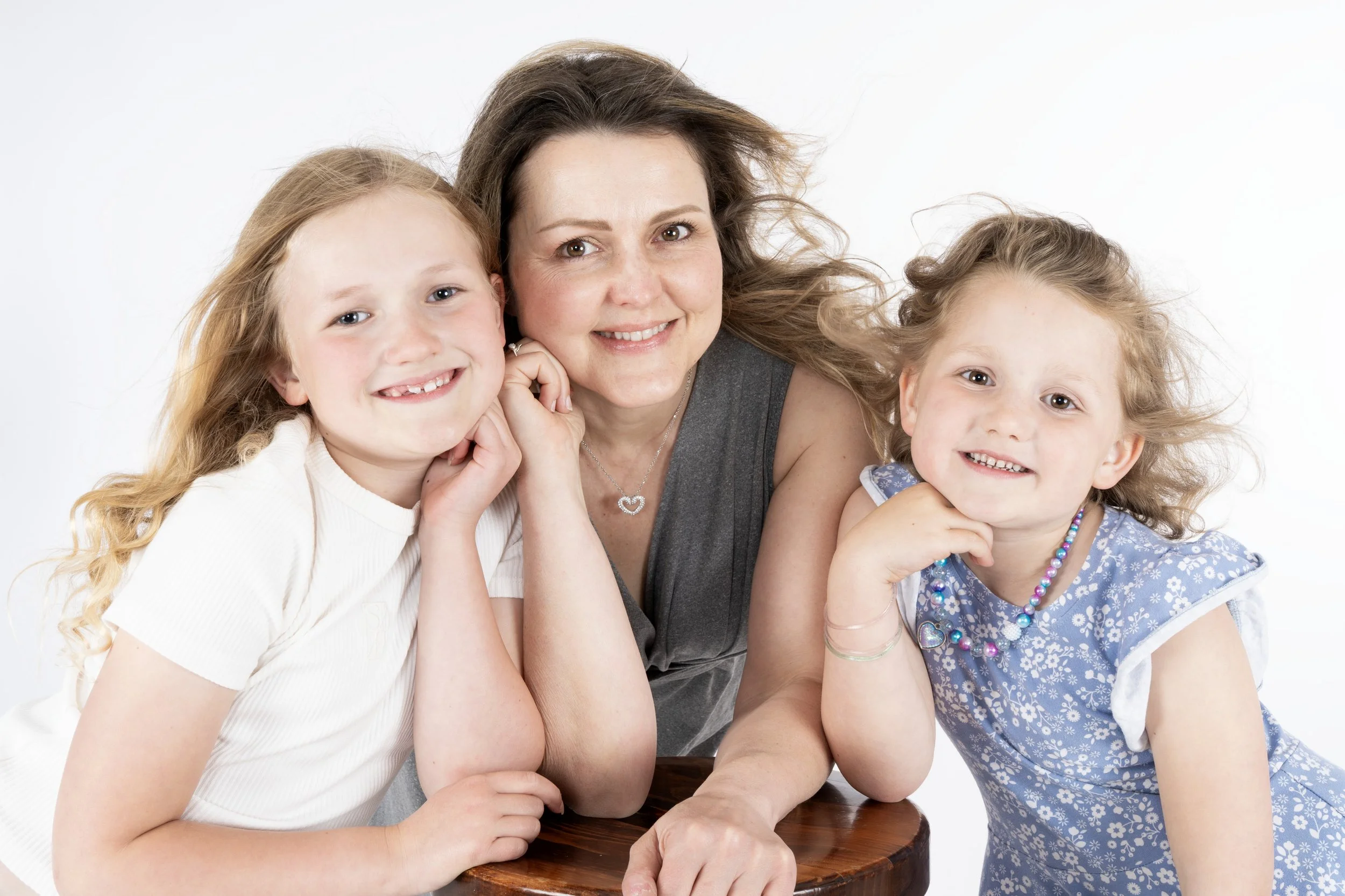 A woman with long brown hair and two young girls with blonde hair smiling and leaning on a table, with a plain white background.