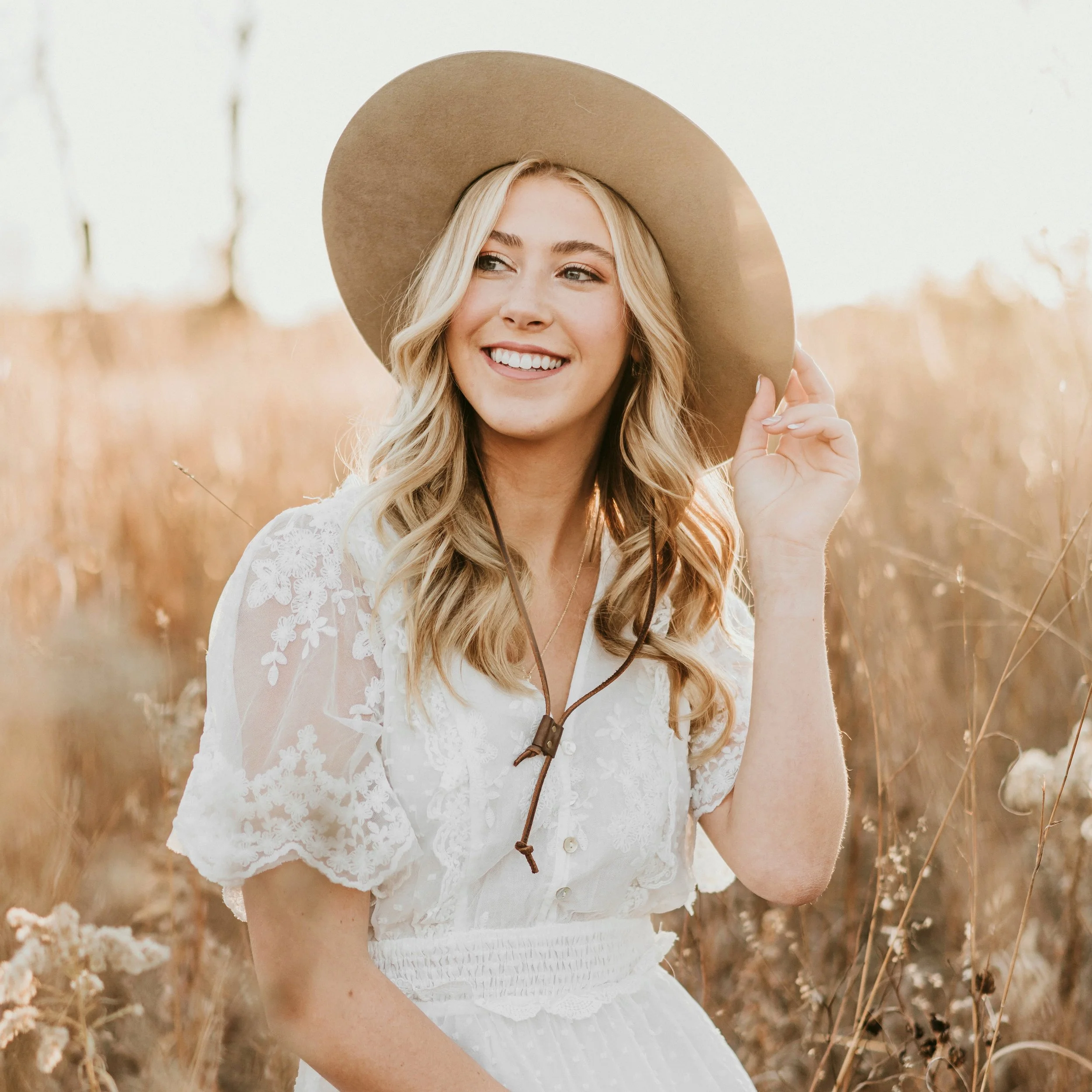 A young woman with long, wavy blonde hair wearing a beige wide-brimmed hat and a white lace top, standing in a field of tall, golden grass during the daytime, smiling and holding the brim of her hat.
