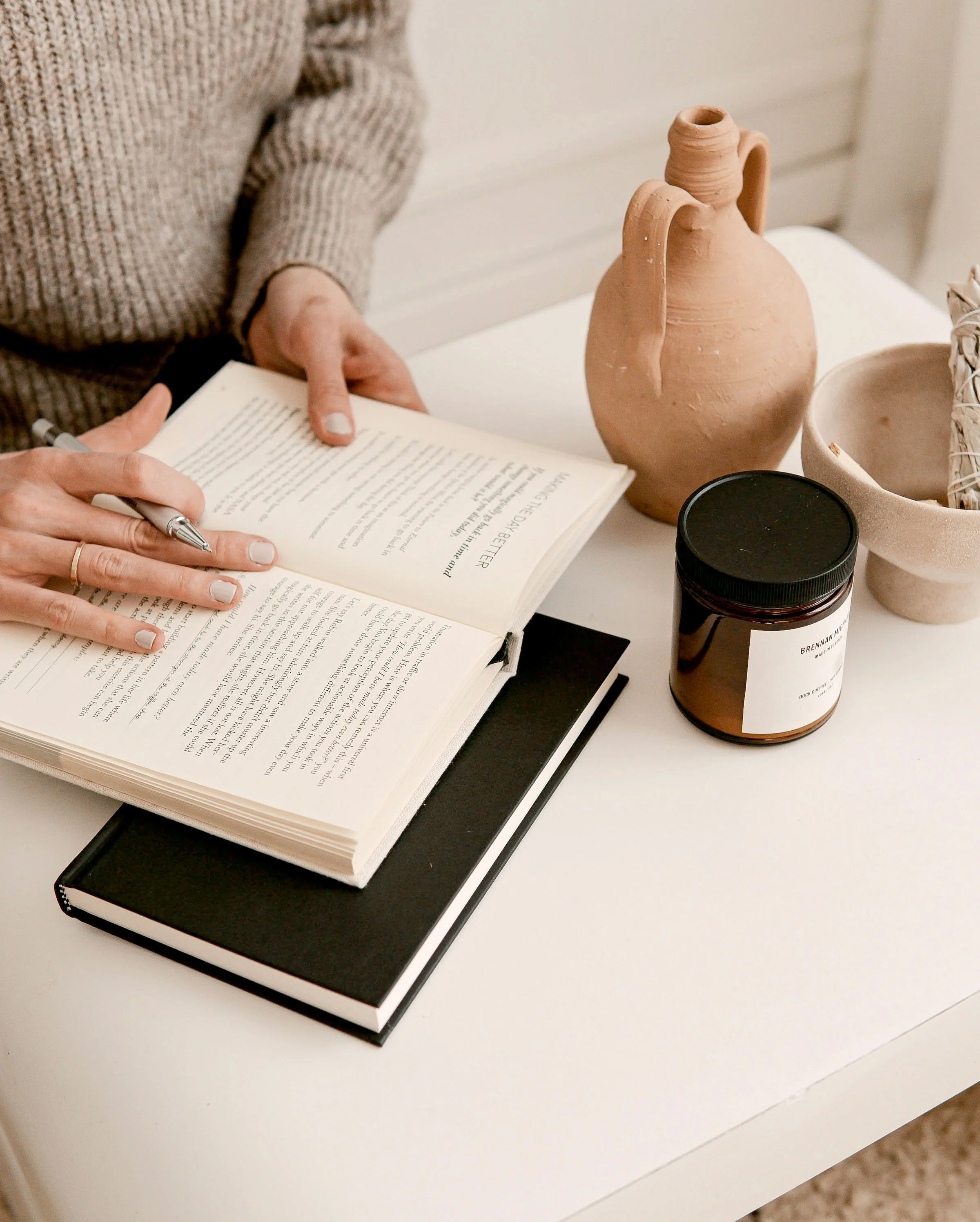 A person wearing a beige knitted sweater is sitting at a white desk, writing in a book with a dental-style mirror and several containers, including a brown jar labeled "BRENNAN MUG," on the desk.