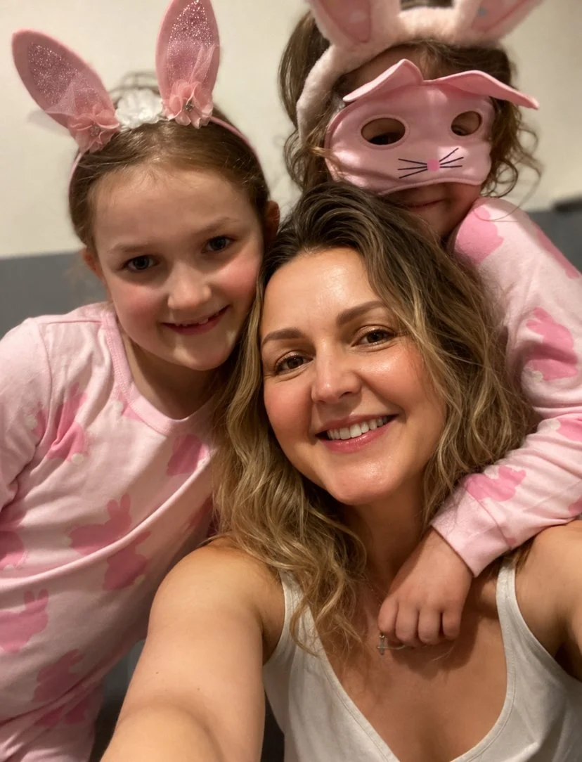 A smiling woman with wavy hair taking a selfie with two young girls dressed in pink, wearing bunny ear headbands and a pink bunny mask with bunny ears and whiskers. All three are celebrating Easter.