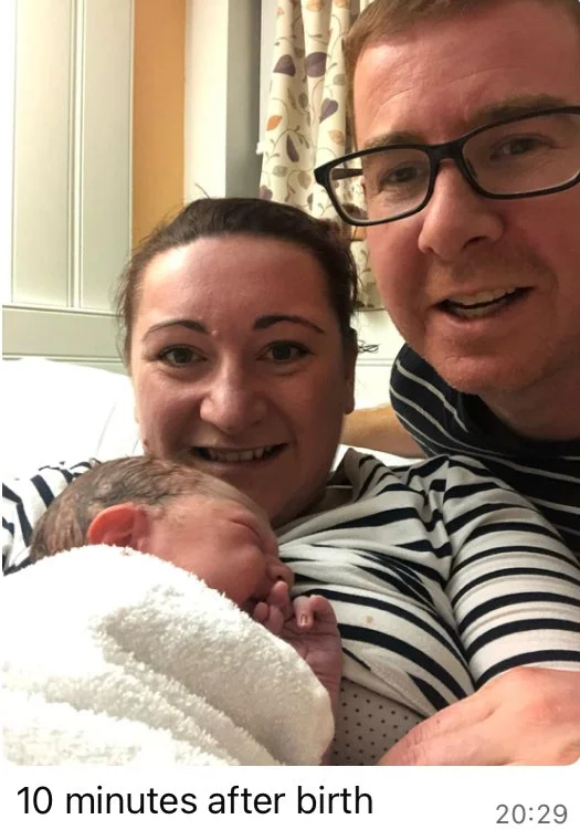 A family photo showing a woman, a man, and a newborn baby in a hospital room. The woman is holding the baby close, and all three are smiling. The woman and man are wearing striped shirts, and there are hospital curtains in the background. The caption reads '10 minutes after birth'.