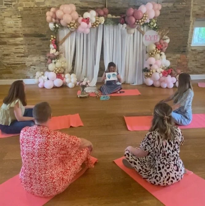 A group of children sitting on pink mats in a room decorated with balloons, listening to a woman reading a book. The balloons are in shades of pink, white, and gold, arranged around a fabric backdrop with a small sign that says 'LYB'.