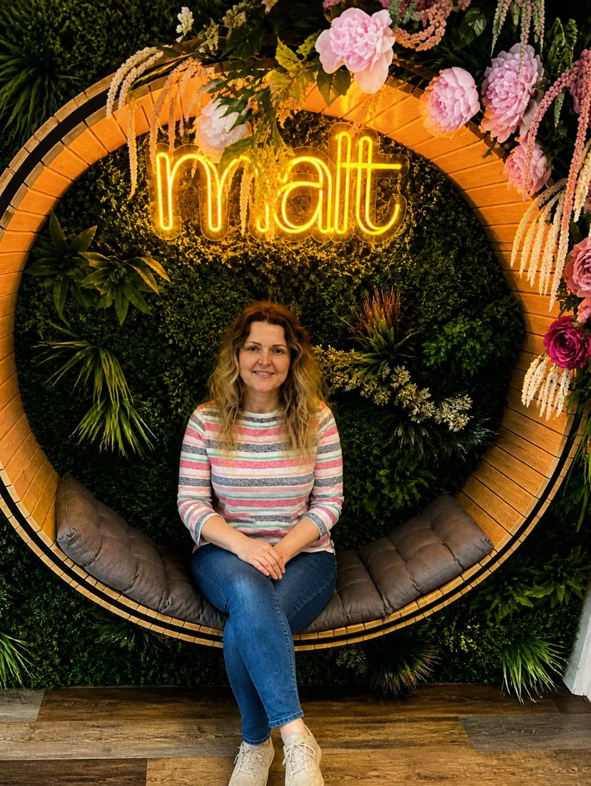 A woman seated on a circular swing with cushions in front of a green leafy wall with pink and purple flowers and a neon sign that reads "malt."