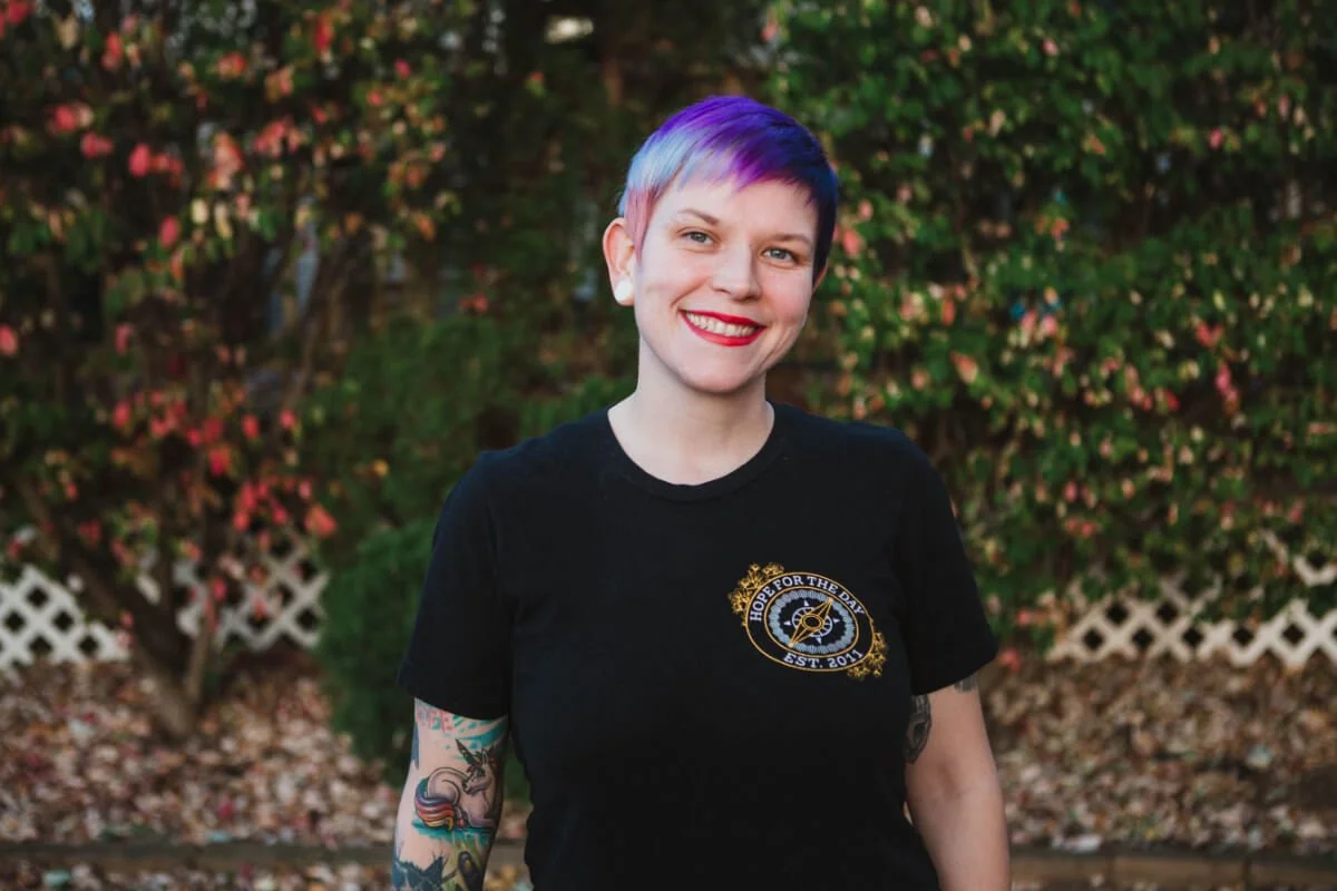 A woman with short purple and blue hair, wearing a black t-shirt with a gold and white logo that reads "Hope for the Day" and features a guitar, outdoors with greenery and pink flowers in the background.