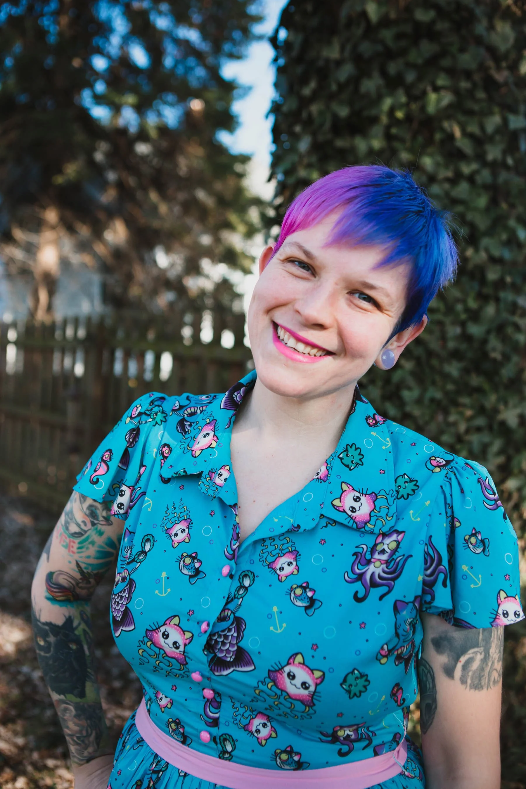 A woman with short, multicolored hair (pink and blue), smiling outdoors with a background of trees and a wooden fence. She is wearing a blue dress with a pattern of unicorns and tattoos on her arms.
