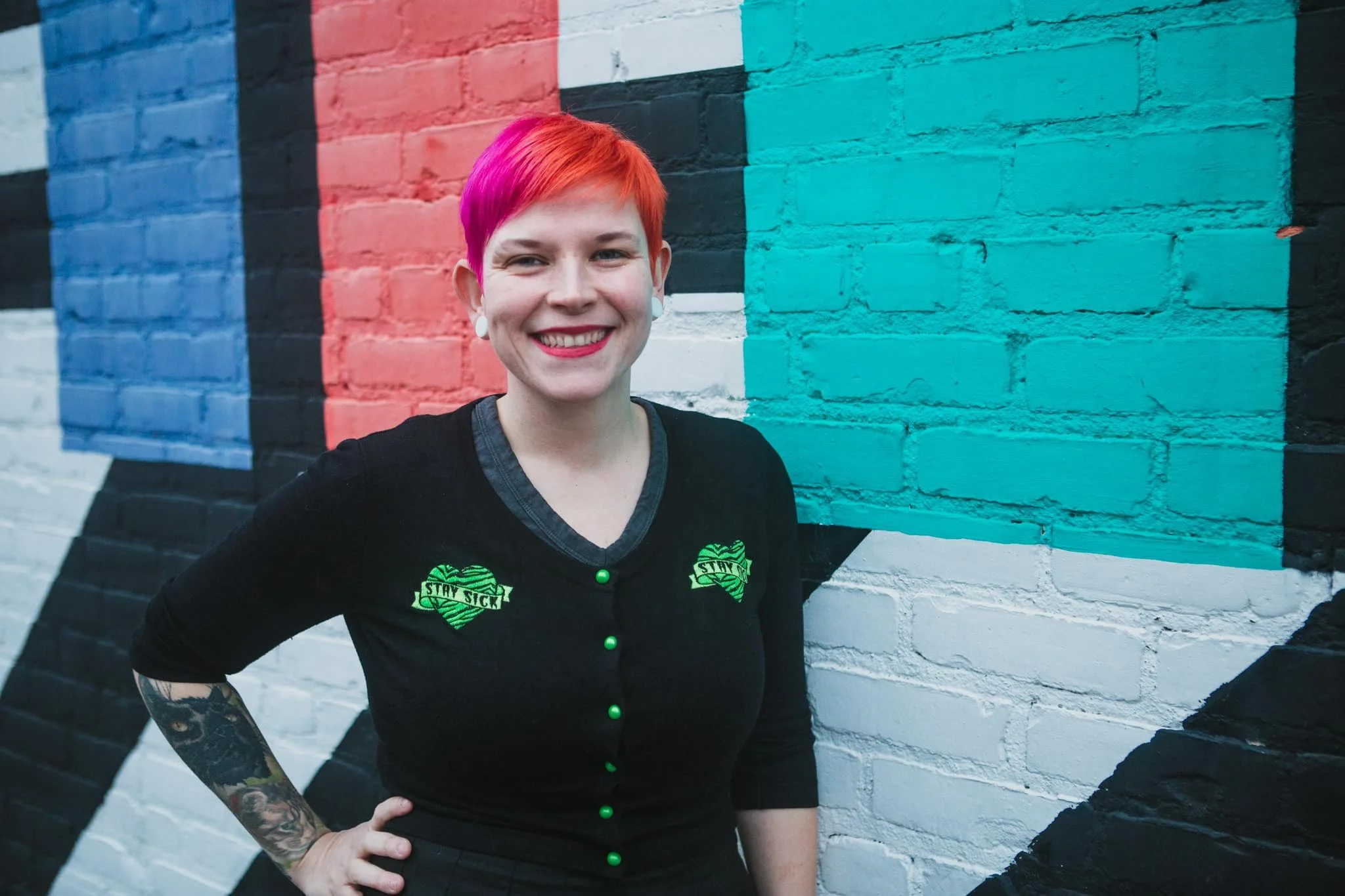 A woman with short red hair and a bright smile standing in front of a colorful mural on a brick wall. She is wearing a black shirt with green buttons and a green and black logo that says "Stay Sick".