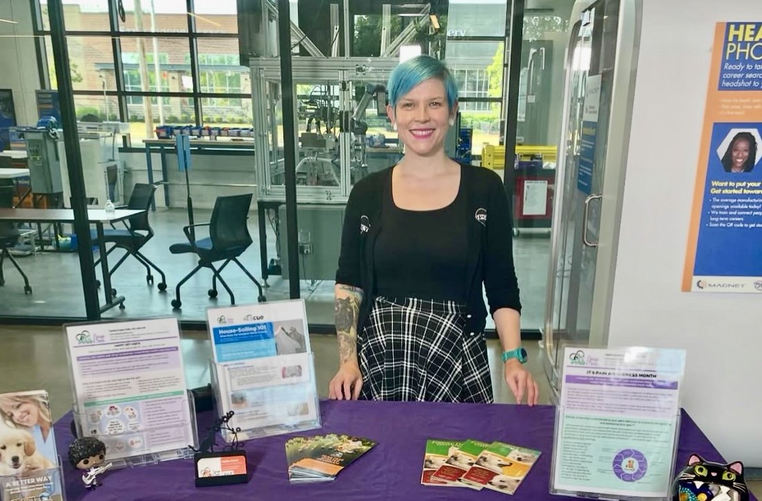 A woman with blue hair standing behind a table with informational materials at an indoor event. The table has brochures, flyers, and small decorative items. She is smiling and wearing a black top with a checkered shirt tied around her waist, in a bright, modern space with chairs and windows.