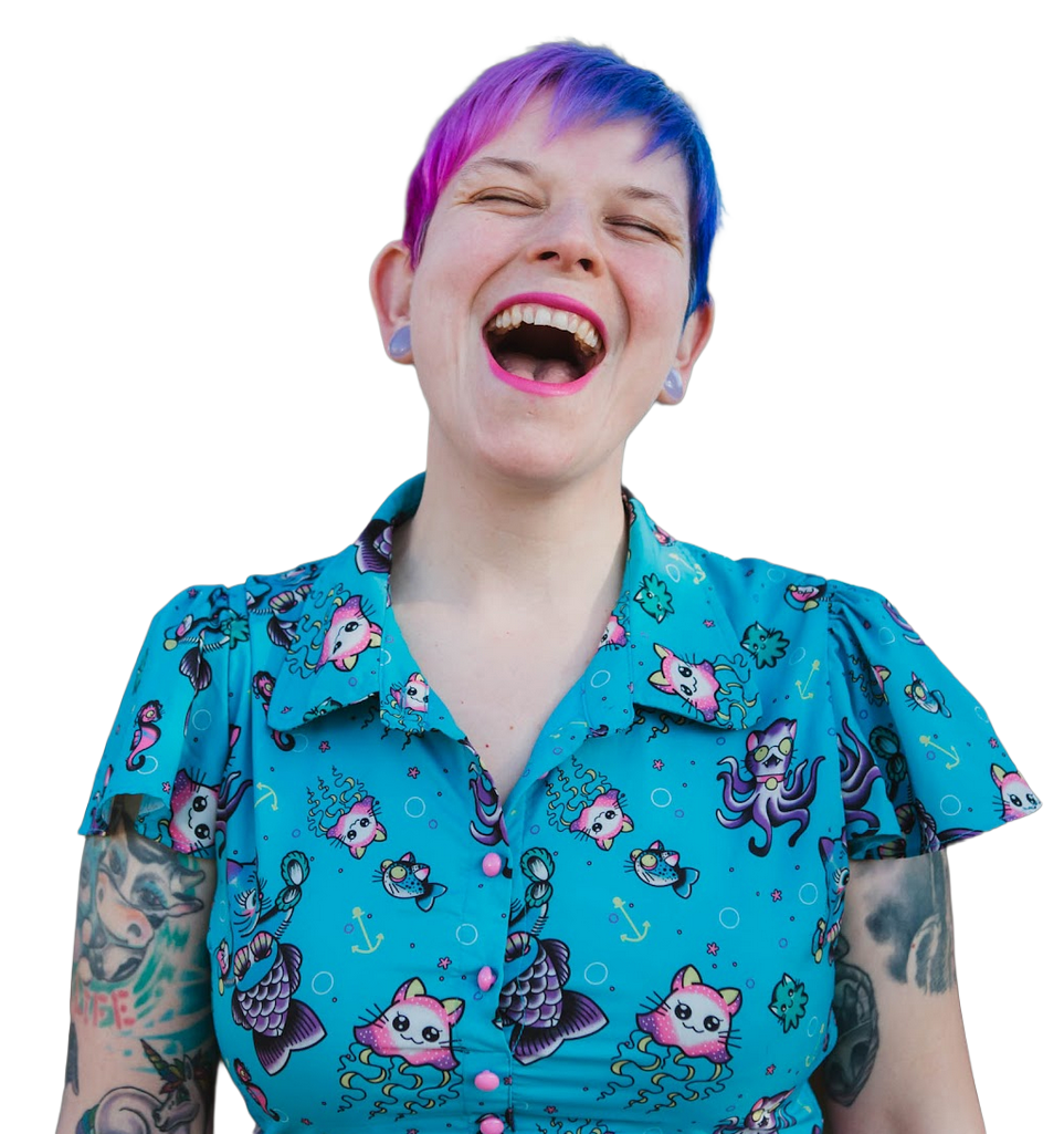 A woman with rainbow-colored short hair, laughing happily. She is wearing a blue shirt with a pattern of cartoon cats, fish, and anchors. She has tattoos on her arms and is wearing earrings and pink lipstick.