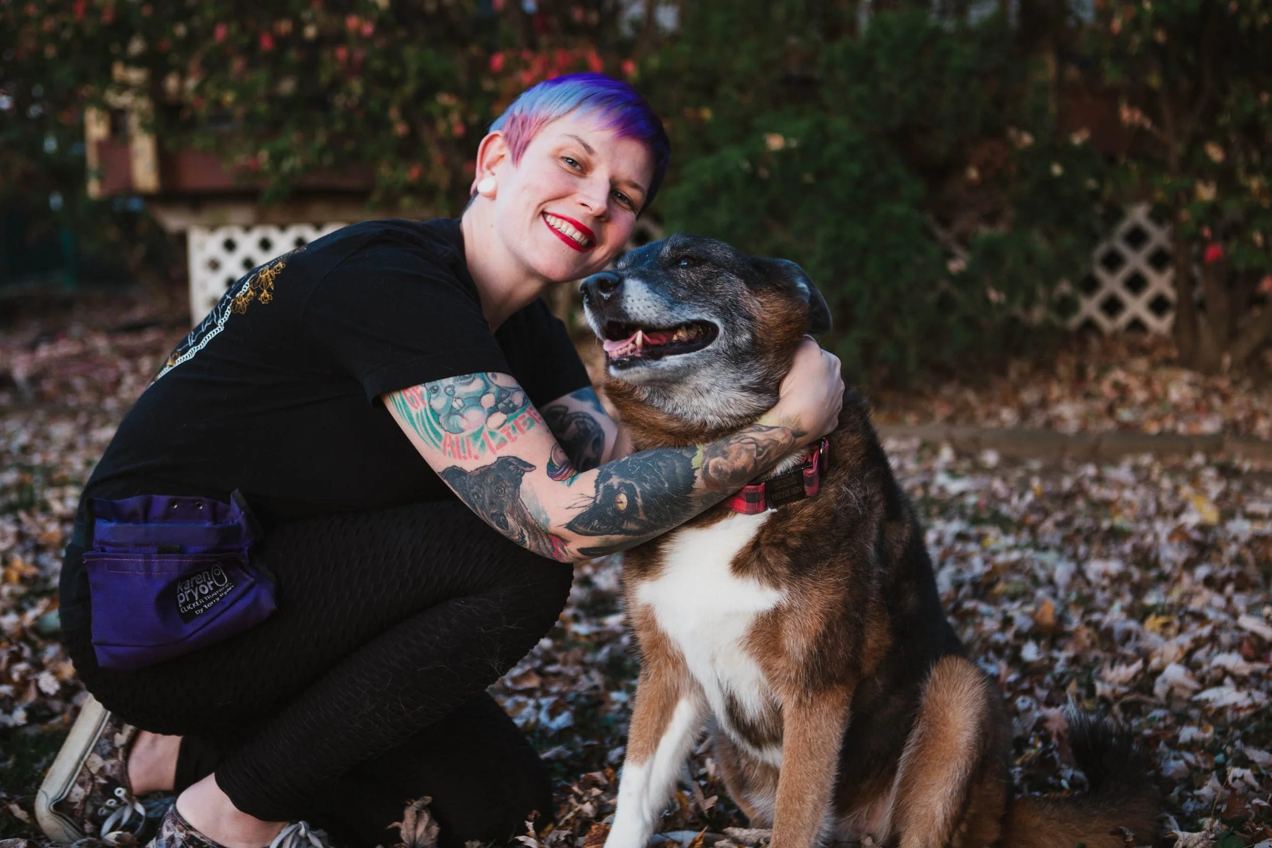 A person with short colorful hair and tattoos hugging a large dog outdoors with a background of trees and fallen leaves.