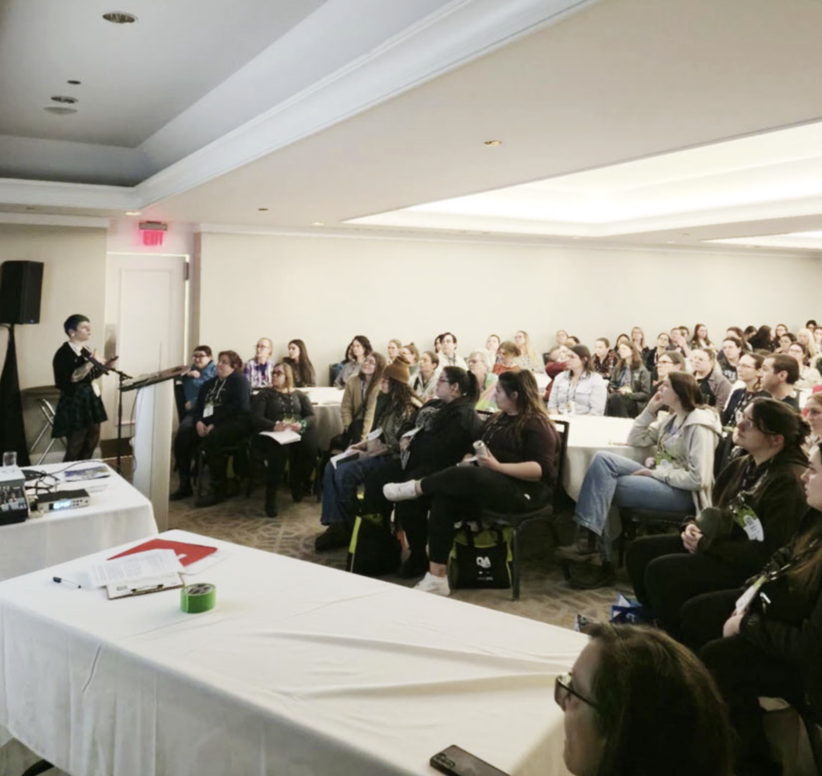 A woman in black clothing and glasses giving a presentation to a seated audience in a conference room with beige walls and ceiling. The audience is listening attentively, and some are taking notes. There are tables with papers and a roll of green tape in the foreground.