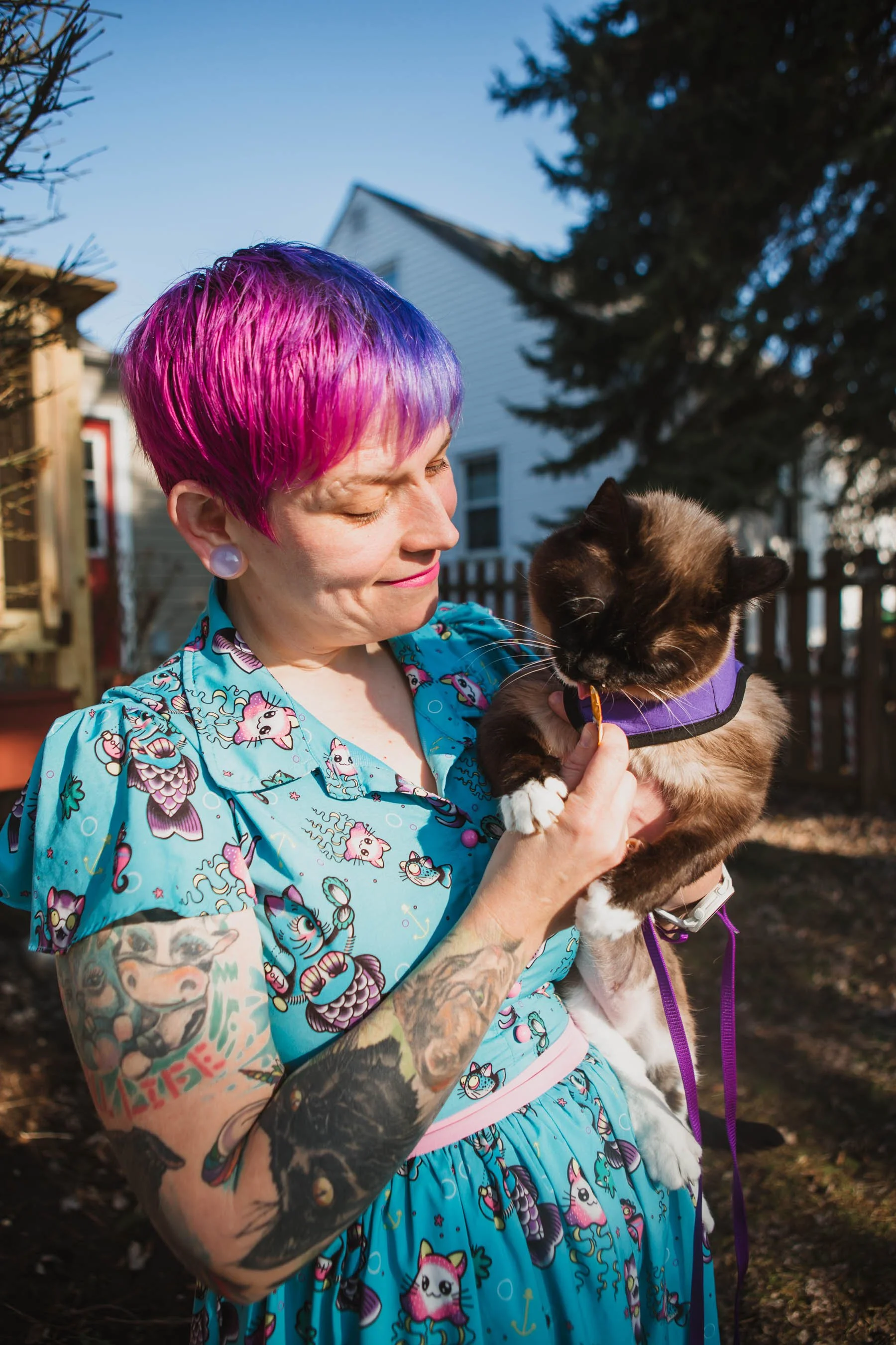 A woman with short, pink and purple hair holds a black and white cat wearing a purple harness outdoors on a sunny day. The woman is smiling with her eyes closed, wearing large white earrings and a colorful, cartoon-themed dress. In the background, there are trees, a white house, and a wooden fence.