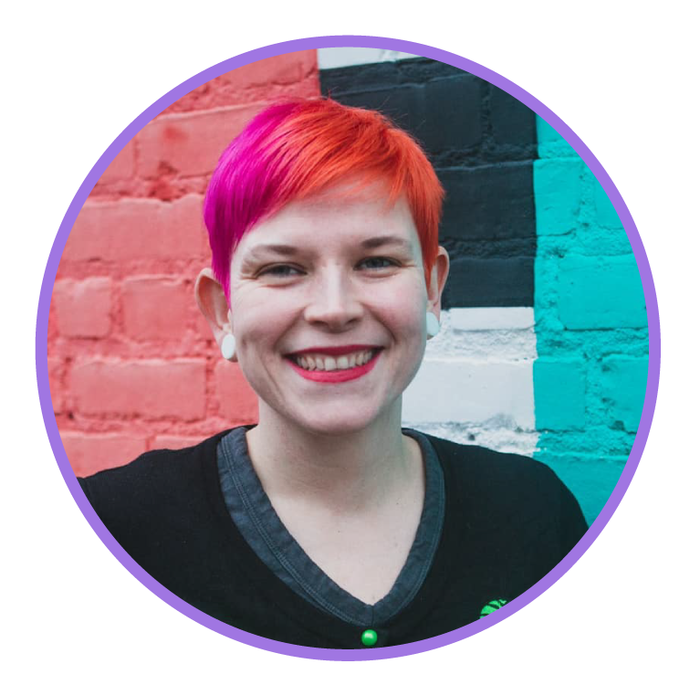 A woman with short, vibrant pink and red hair smiling, standing in front of a colorful painted brick wall.
