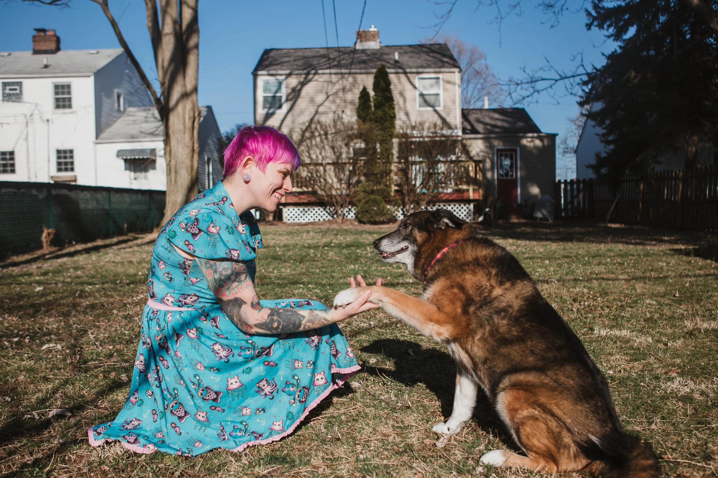 A woman with pink and purple hair sitting on grass, smiling, as she shakes hands with her dog in a backyard with houses in the background.