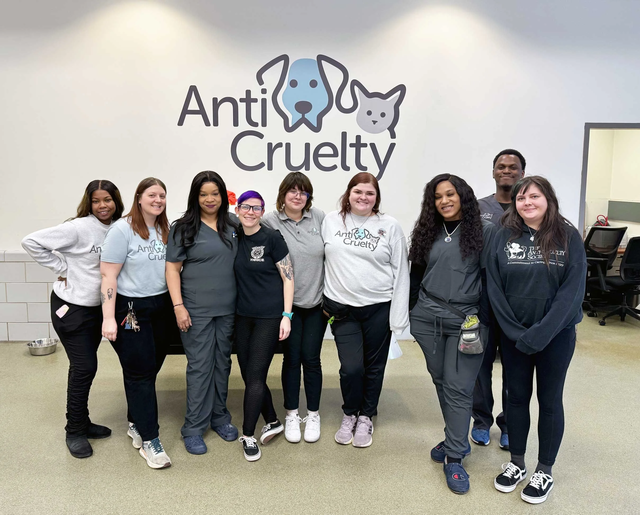 Group of diverse people standing together in front of a wall with a sign that reads 'Anti Cruelty' and features images of a dog and a cat, smiling and posing for the photo.