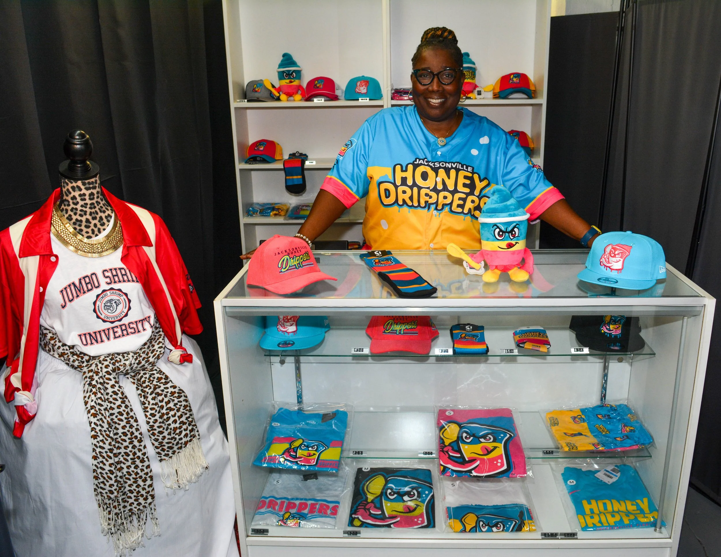 A woman wearing glasses and a colorful shirt stands behind a display counter with various merchandise, including hats, shirts, and a plush toy, all branded with 'Honey Drippers.' Behind her are white shelves filled with more hats and merchandise, and to her left is a mannequin dressed in a red and white jacket with leopard print fabric and a gold necklace.