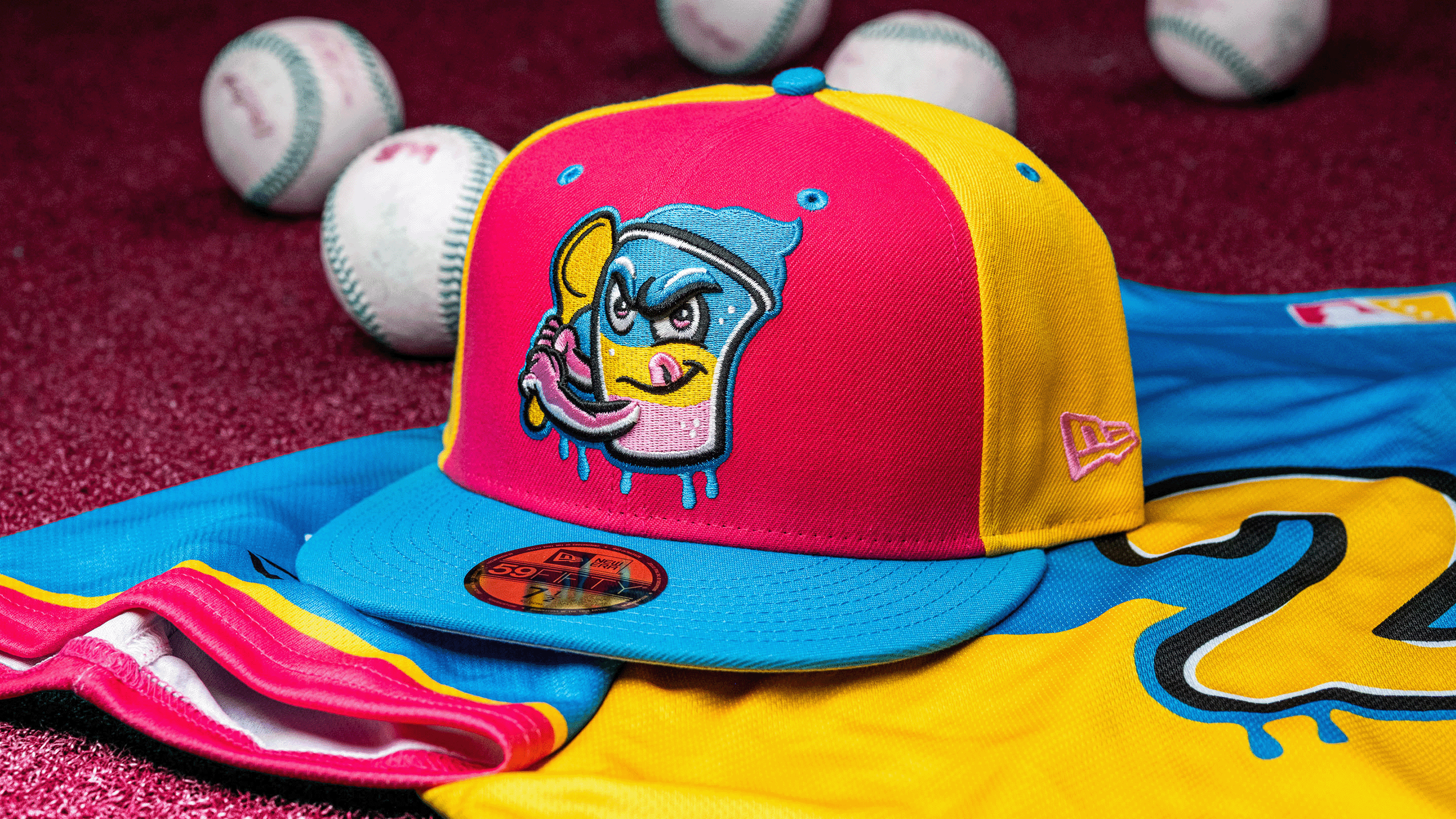 Colorful baseball cap with cartoon character embroidery, surrounded by stacked white baseballs with pink and blue stitching, and partially folded yellow and blue clothing on a textured red surface.