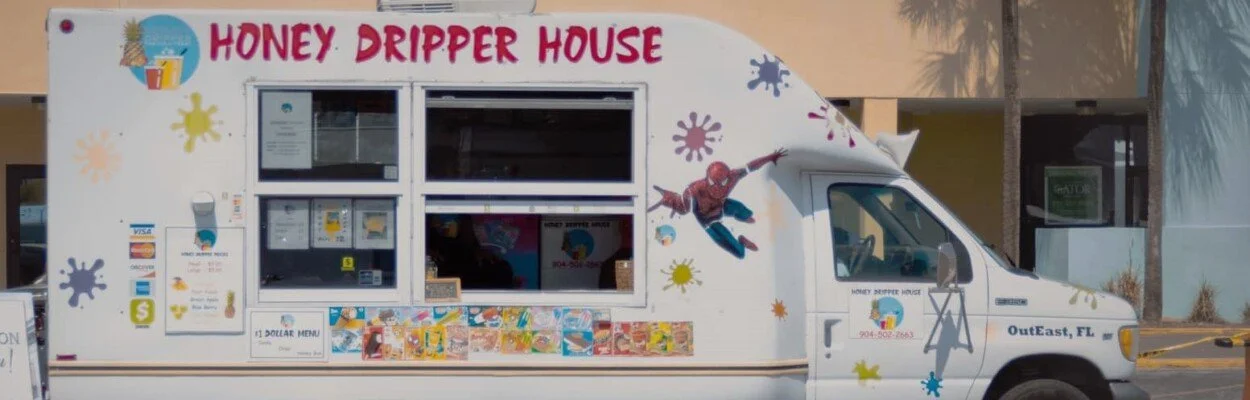 A white food truck named 'Honey Dripper House' in OutEast, Florida, decorated with colorful paint splatters and a Spiderman decal, featuring a serving window and menu signs.