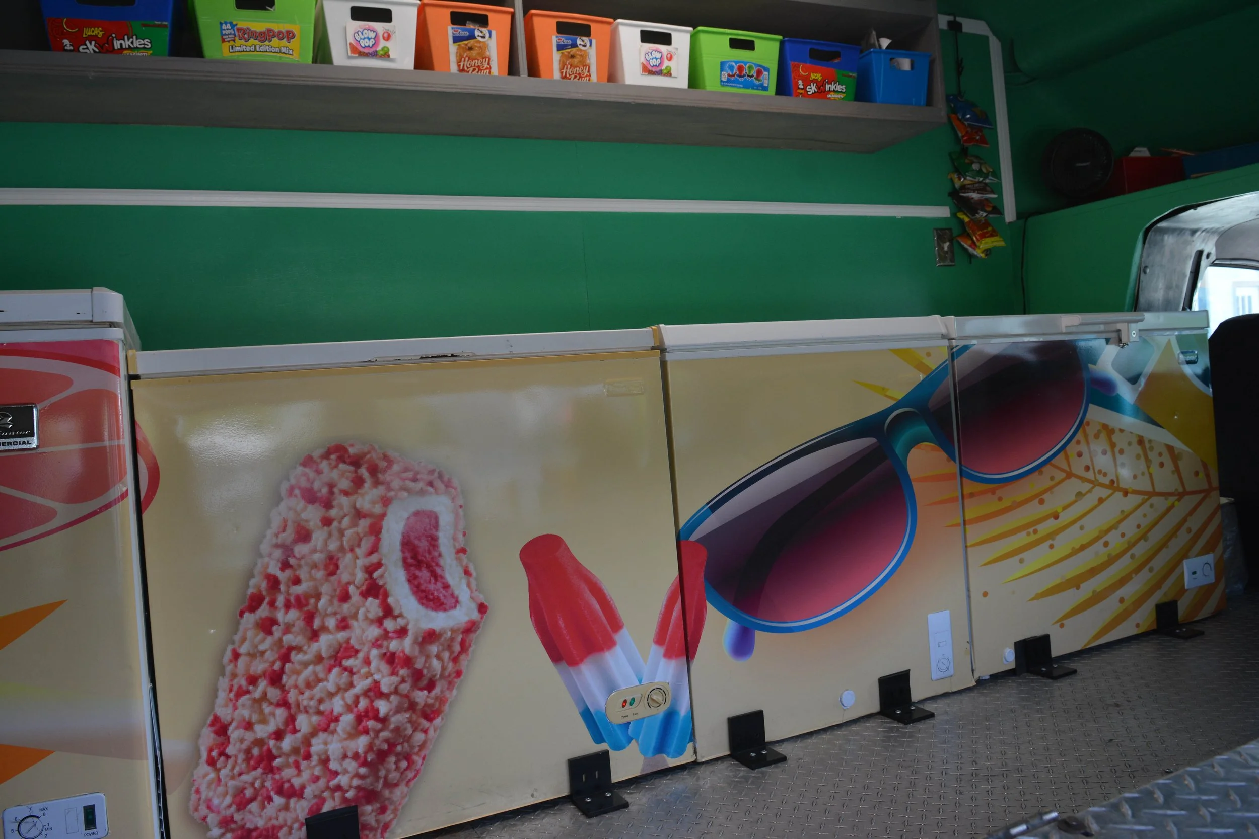 A row of colorful ice cream vending machines with images of ice cream treats and popsicles on them, set against a green wall with snack shelves above.