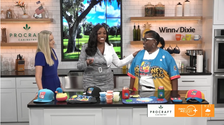 Three women are in a kitchen studio set, engaging in conversation and smiling. The kitchen has a large screen in the background showing a park scene, and shelves with decor and kitchen items. The table in front of them has colorful hats, cups, and food items. The setting appears to be a television segment about cooking or crafts.