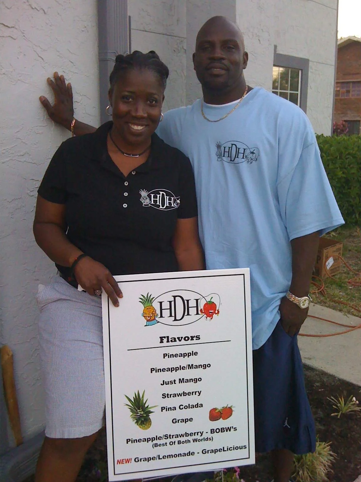 Two people standing outside, holding a sign with various fruit flavors, including pineapple, mango, strawberry, and grape, with a woman on the left wearing a black shirt and a man on the right wearing a light blue shirt.