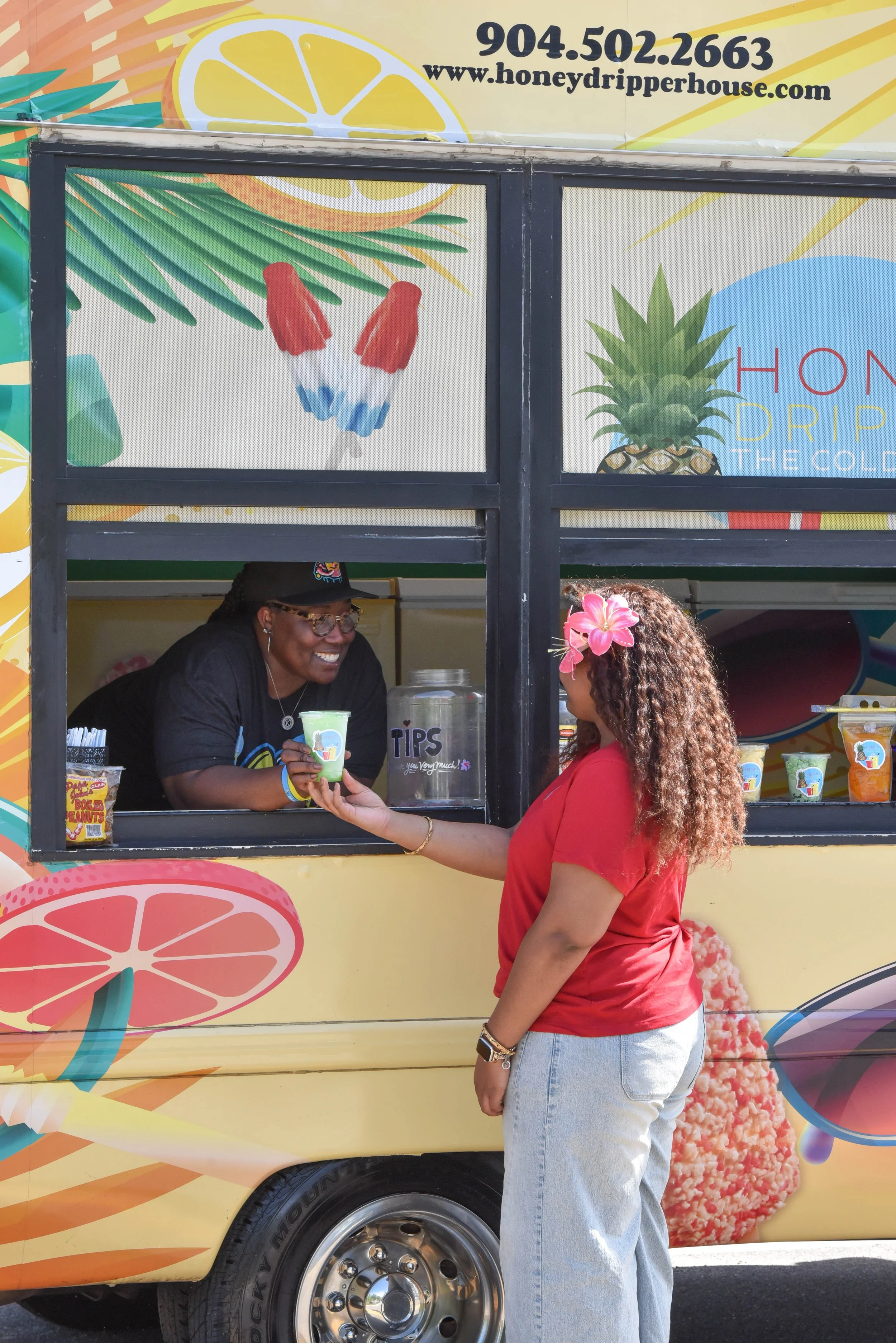 A woman with a pink flower in her curly hair is receiving a cup of watermelon flavored shaved ice or frozen treat from a smiling vendor behind a colorful ice truck decorated with fruit images.