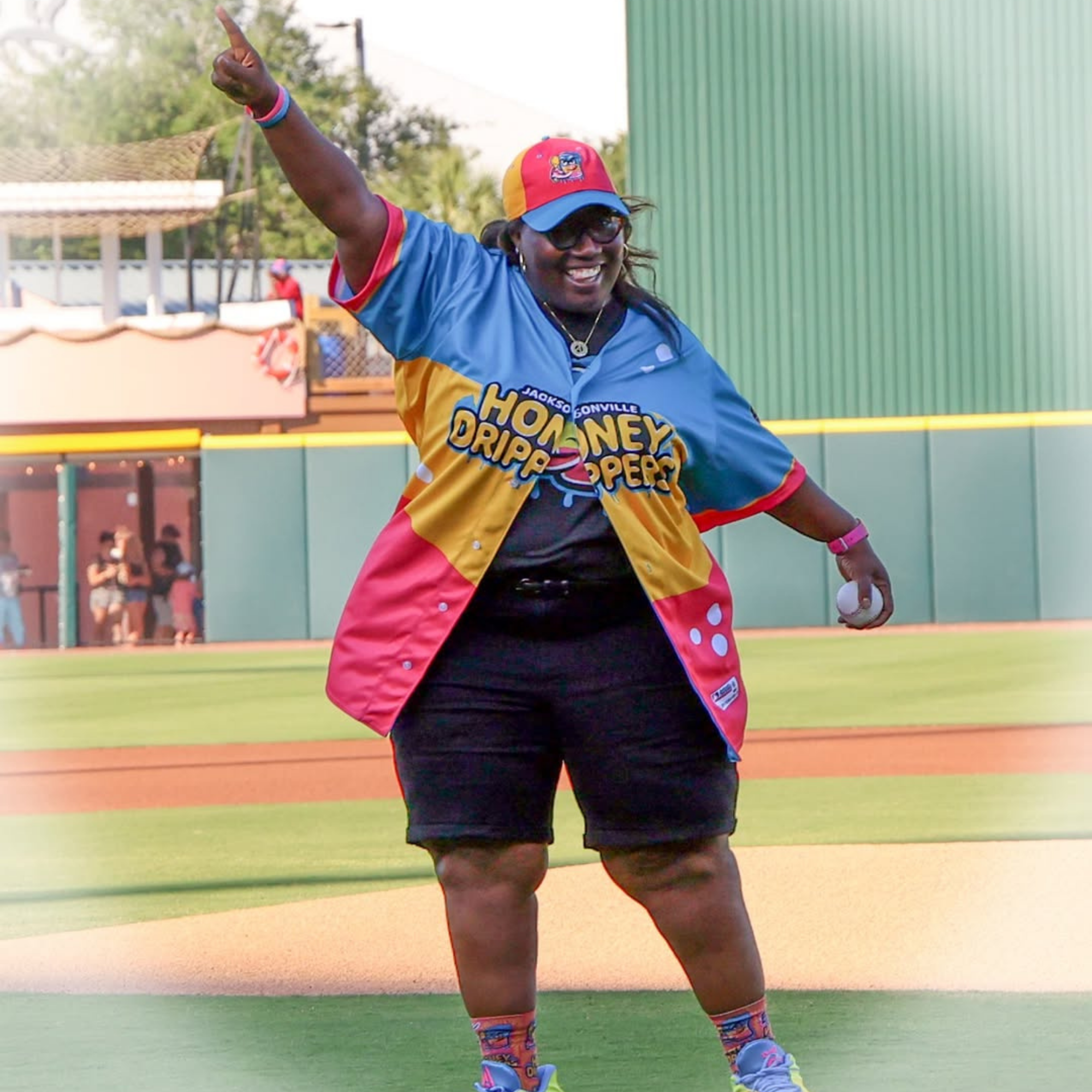 Woman wearing sports attire, a colorful cap, and sunglasses dancing or celebrating on a baseball or softball field, holding a ball in her right hand.