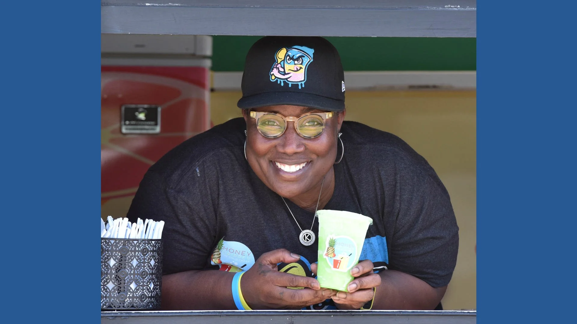 A smiling person wearing glasses and a black cap with a colorful cartoon character, holding a green cup with tropical fruit and pineapple graphics, sitting at a counter with straws.
