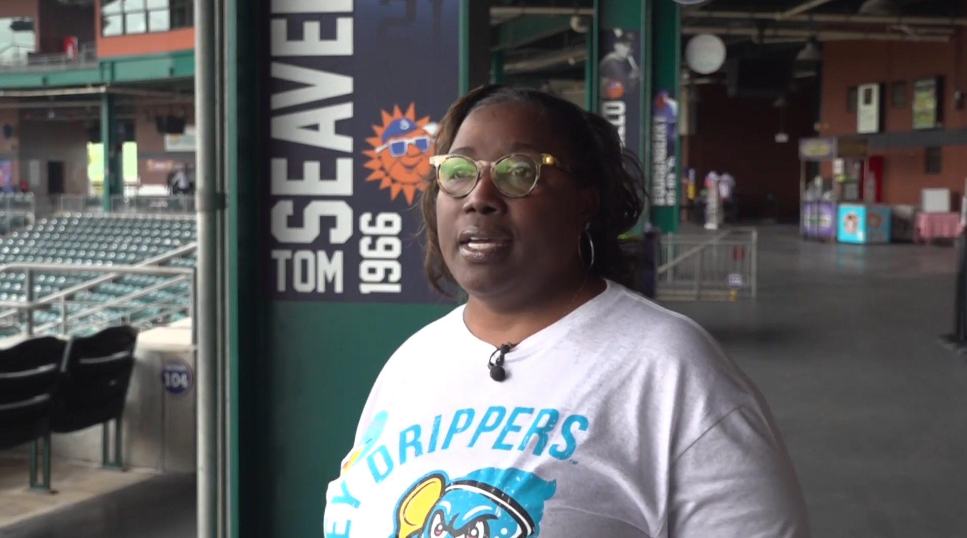 A woman wearing glasses and a white T-shirt with the California Padres logo stands outside a stadium entrance near a sign that reads "Tom Seaver 1966" with a sun and sunglasses graphic.