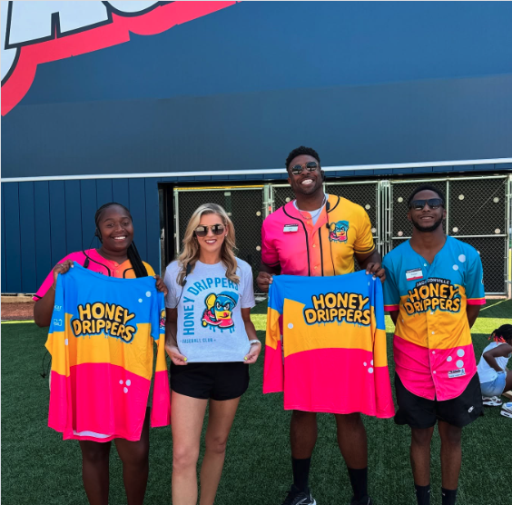 Four people standing in front of a sports stadium holding up jerseys that say "Honey Drippers." The group includes two women and two men, all smiling and wearing colorful uniforms or casual clothing.
