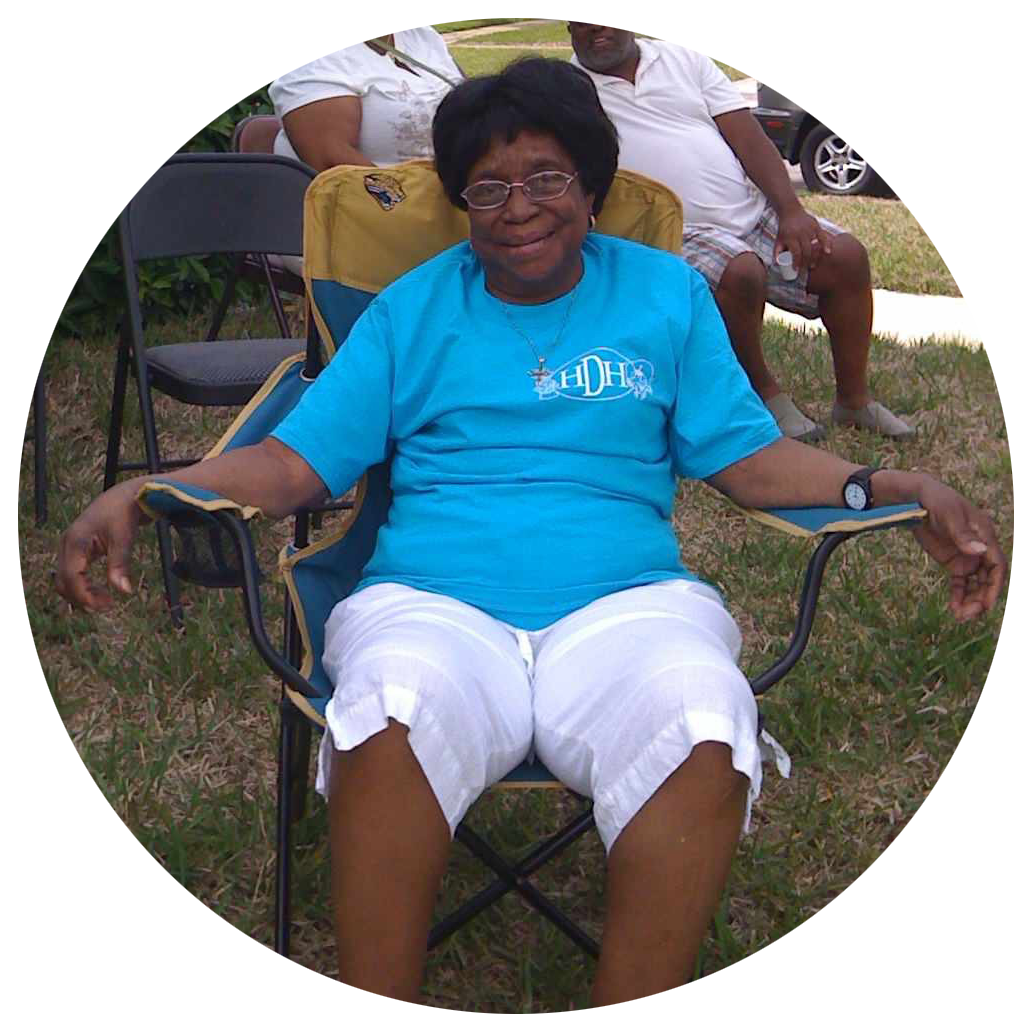 A woman sitting in a foldable lawn chair outdoors, wearing a blue t-shirt and white shorts. She is smiling and wearing glasses. Two people sit behind her on the grass in a park-like setting.