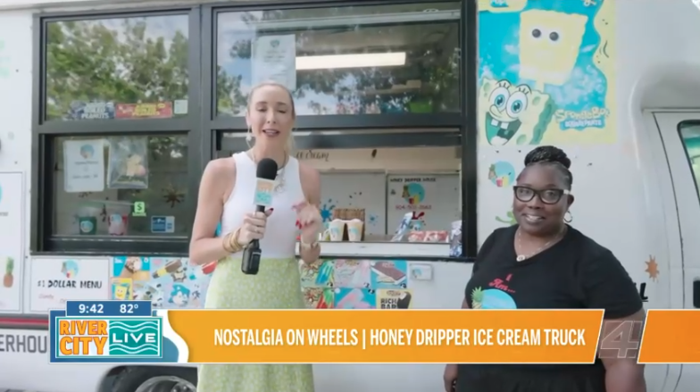 A news reporter holds a microphone, standing outside an ice cream truck with two women, one wearing a black shirt and glasses, and the other in a white top and green skirt.