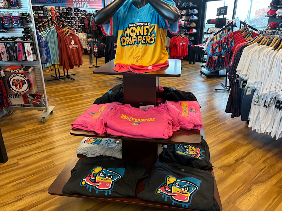 Display table with Jacksonville Honey Drippers merchandise, including yellow and pink T-shirts and black T-shirts with a colorful mascot design, in a retail store with clothing racks and athletic apparel.