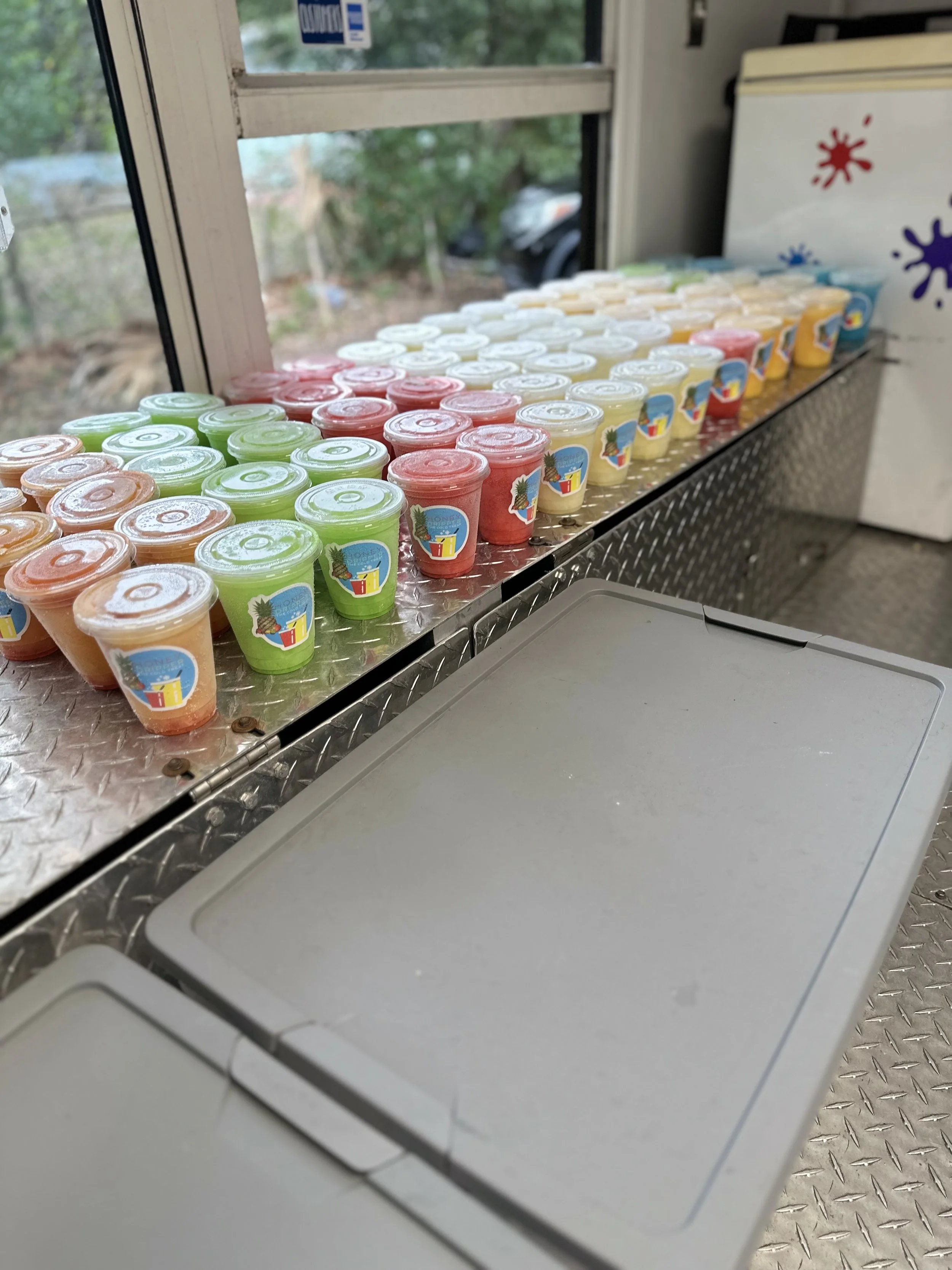 Display of colorful frozen fruit cups with lids on a metal counter next to a window, with a fridge in the background.