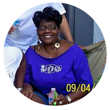 Smiling woman in a purple shirt with glasses and earrings, sitting at a table with water bottles around her.