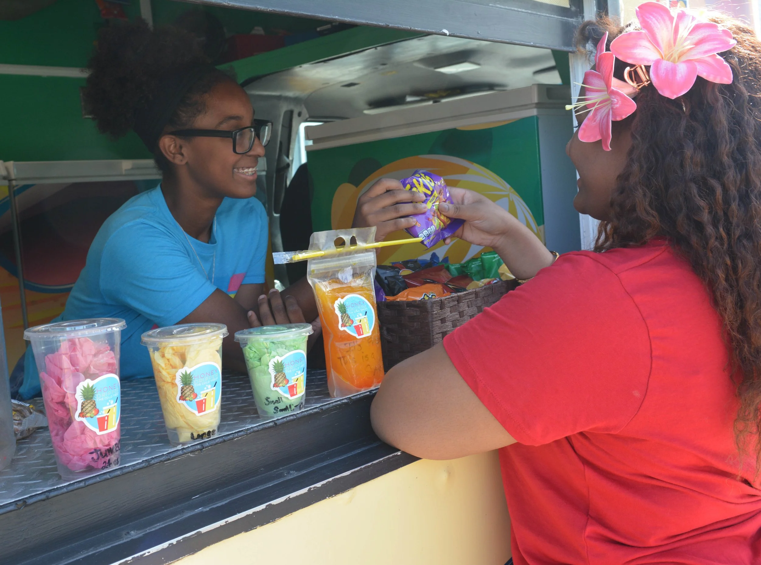A woman with curly hair and a flower headband is purchasing snacks from a vendor at a street stand, with colorful drinks and snacks on display.