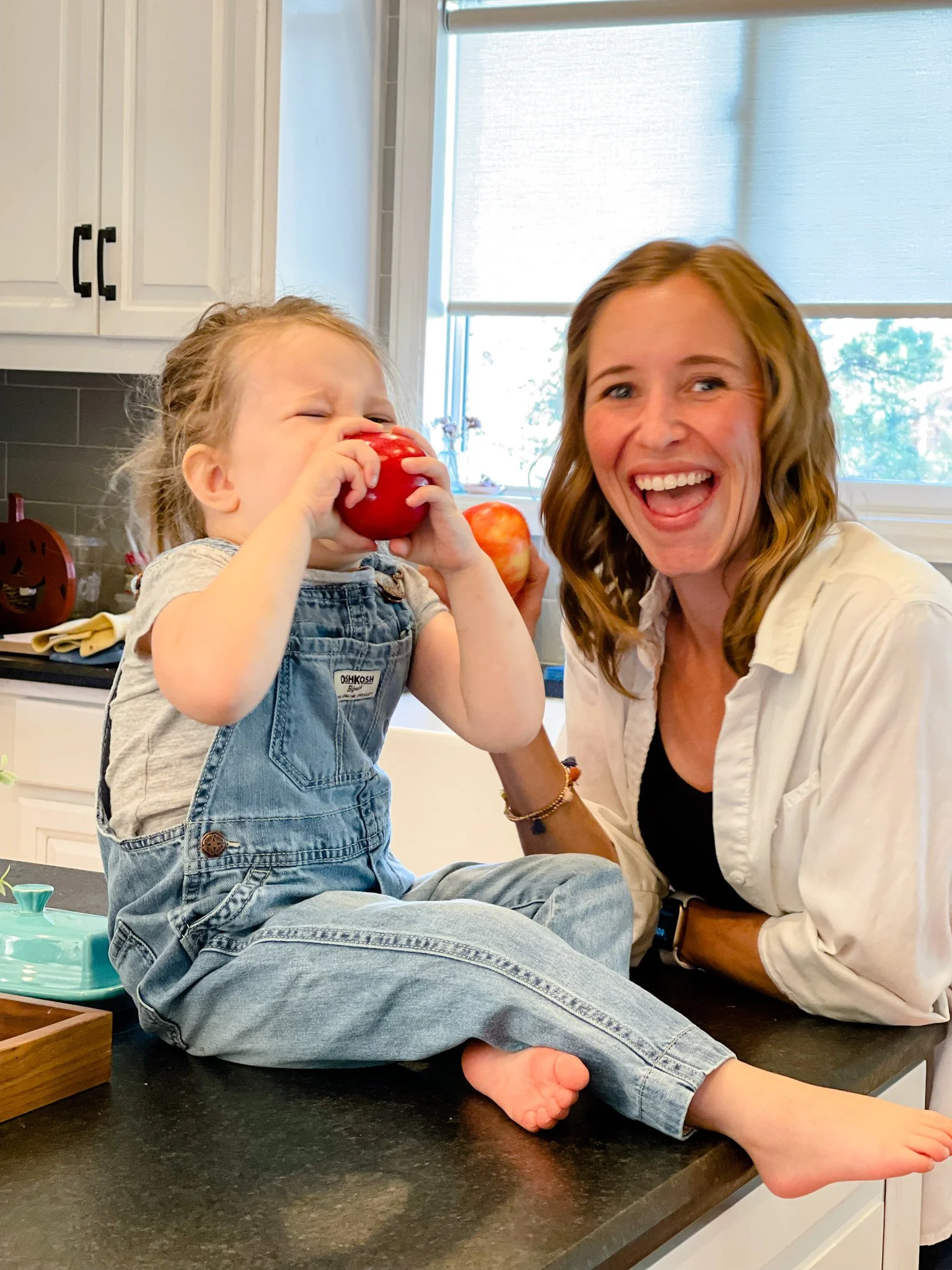 A young child with a toddler's physique, sitting on a kitchen counter, wearing denim overalls and a grey shirt, drinking from a large red apple. An adult woman with shoulder-length wavy reddish-brown hair and light skin, smiling broadly, leaning close to the child, with her mouth open. The background shows a kitchen with white cabinets, a window with closed blinds, and some kitchen items.
