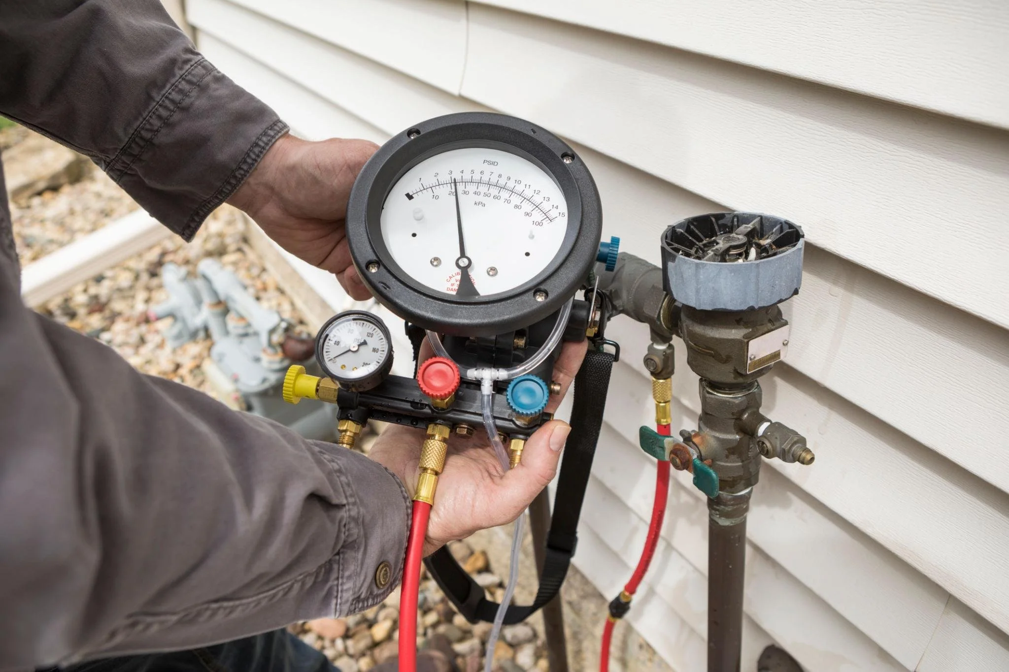 A person is using a manifold gauge set to measure refrigerant pressure on an outdoor air conditioning unit next to a house with white siding.