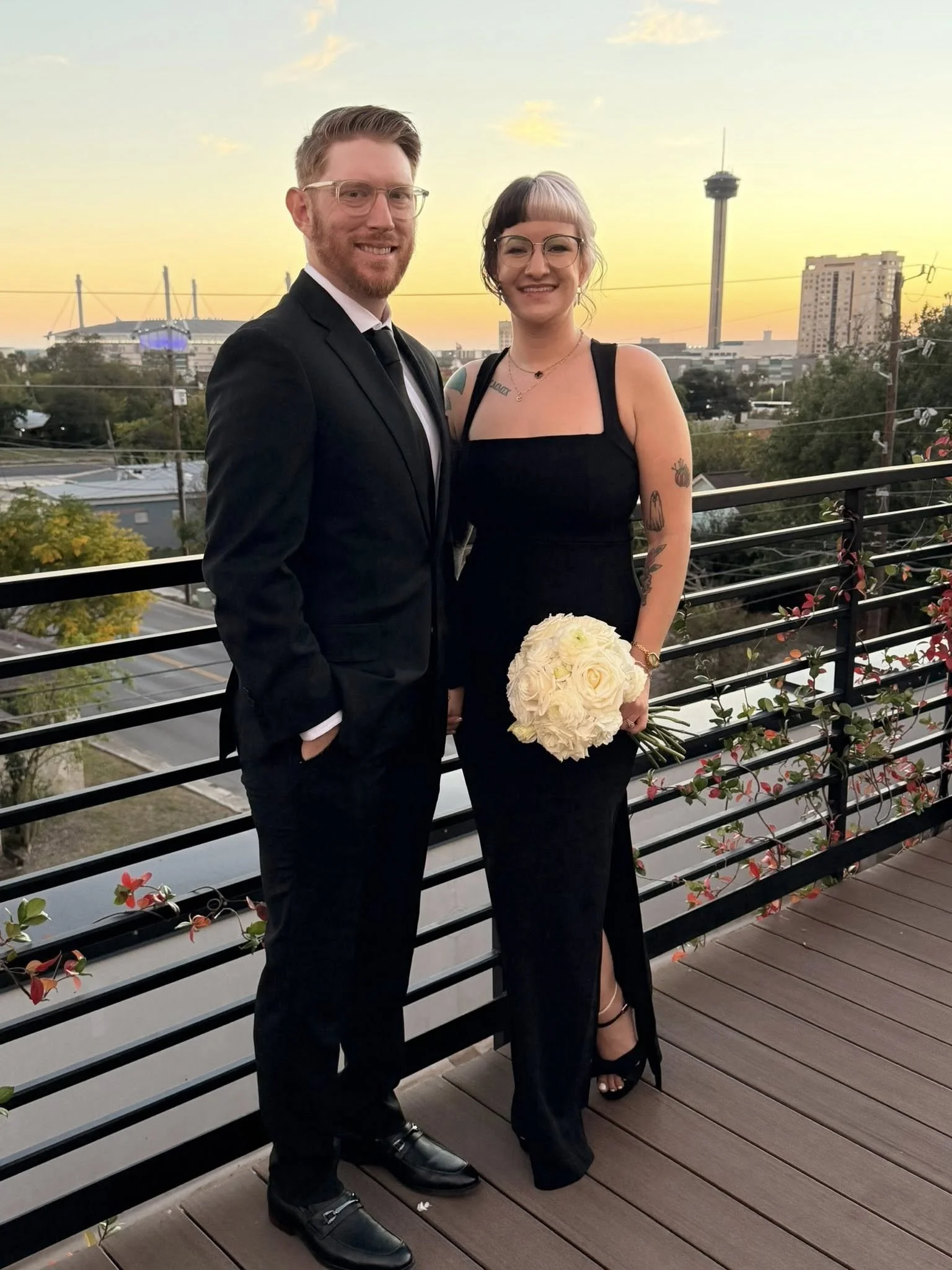 A couple dressed in formal attire standing on a balcony at sunset, with a city skyline and a roller stadium in the background. The woman is holding a bouquet of white roses.