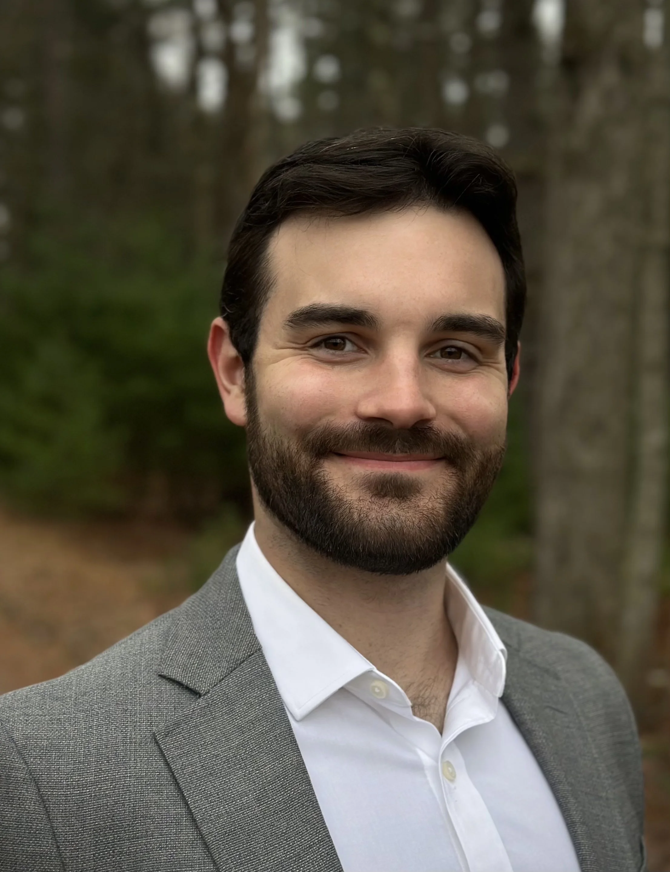 Close-up of a smiling man with dark hair and beard wearing a gray suit and white shirt outdoors.