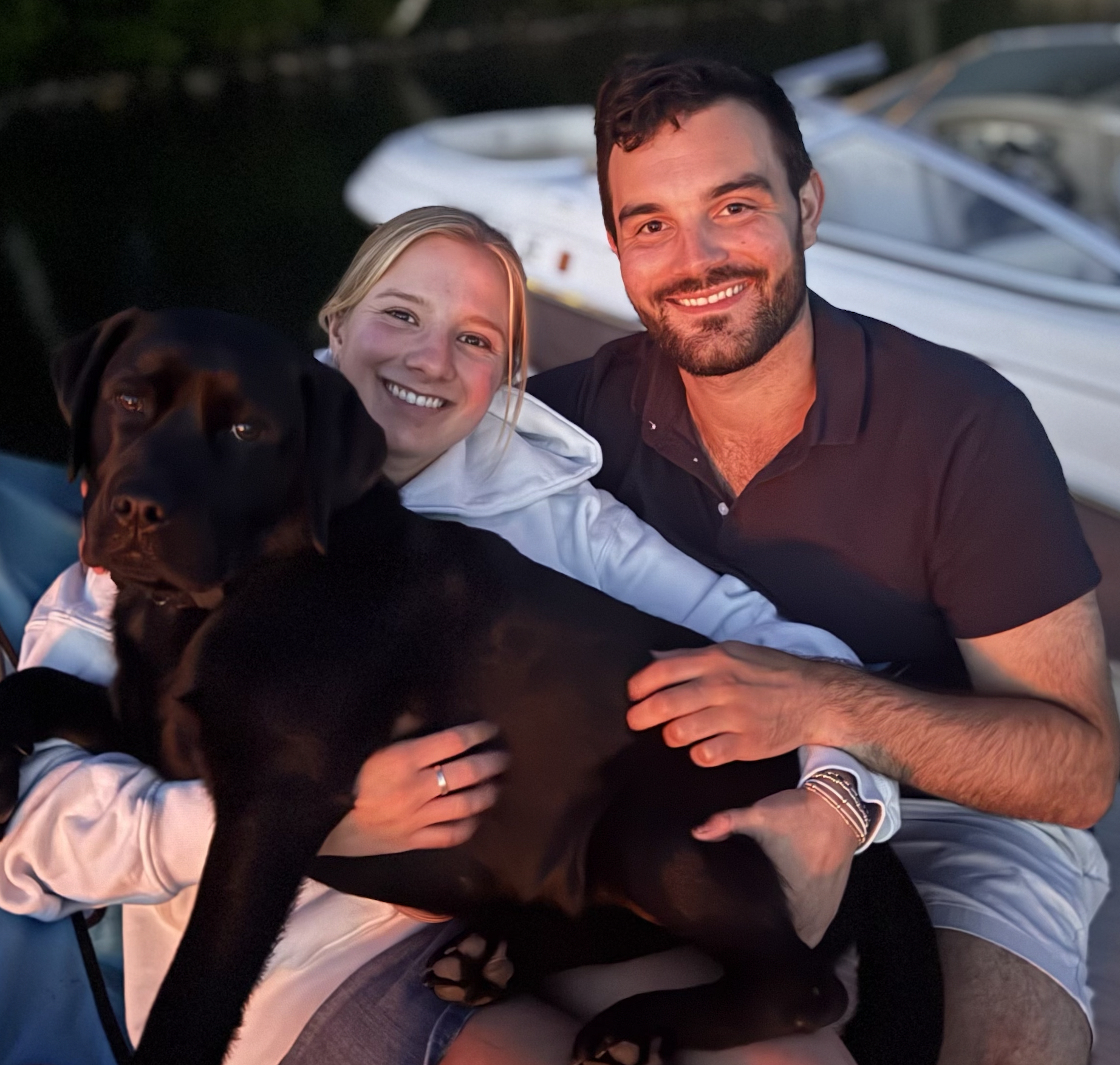 Two adults smiling, hugging a black dog in an outdoor setting near water, during sunset.